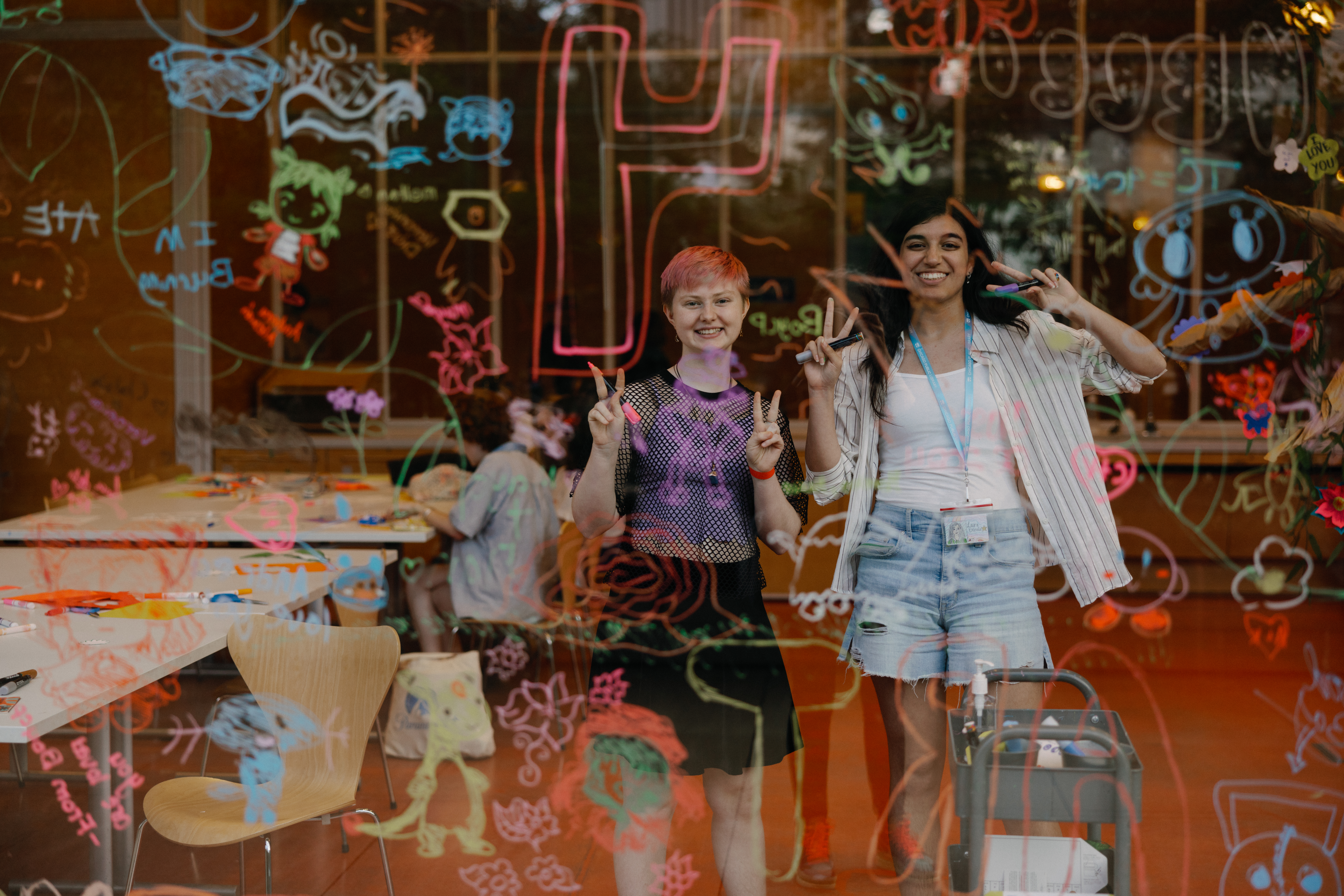 Two teens—a shorter, light-skinned teen with cropped hair and a taller, medium-skinned teen with long hair—photographed through a large glass window with neon writing and doodles all over it. They flash peace signs with their hands and smile.
