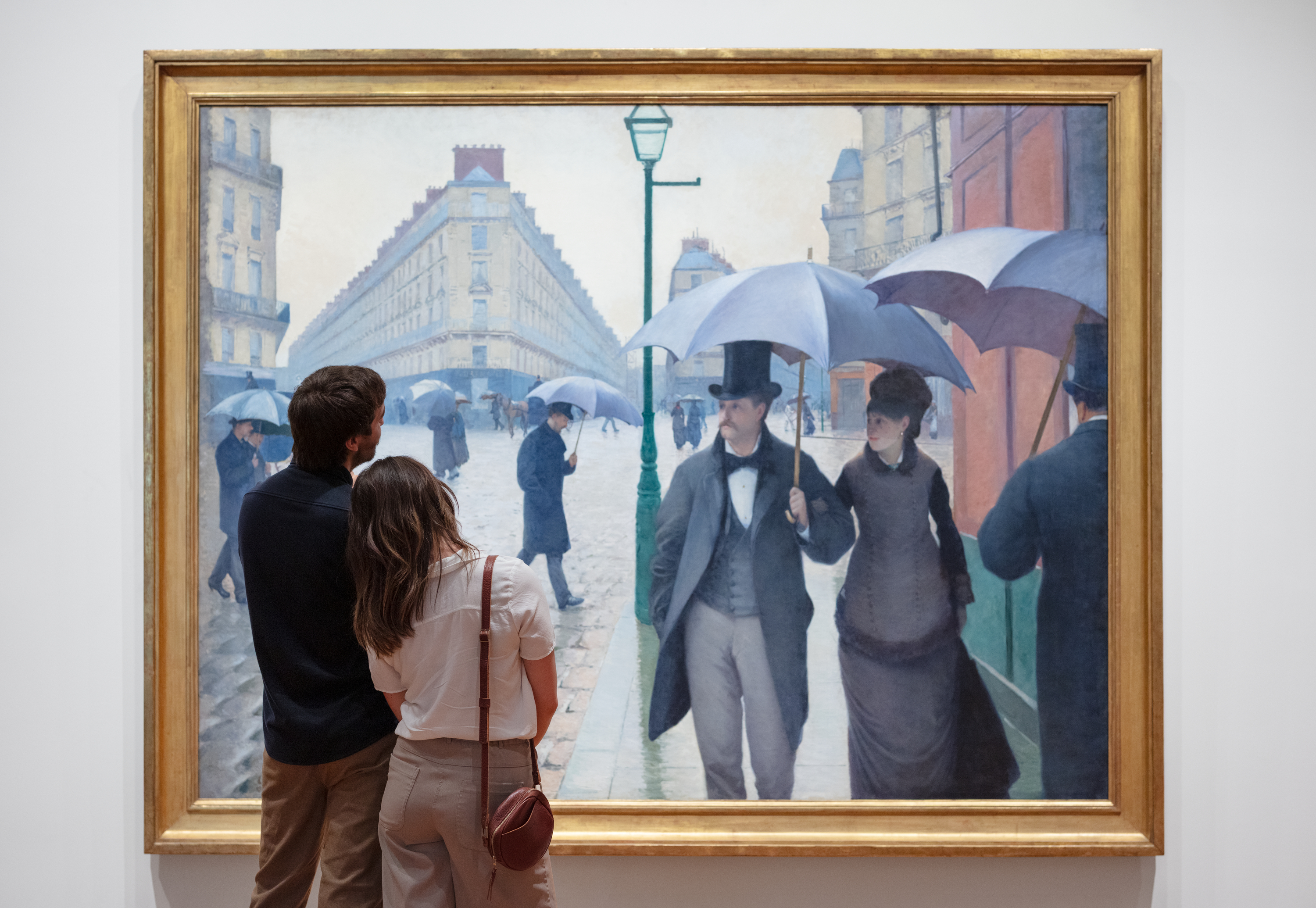 A couple stands before Gustave Caillebotte’s painting “Paris Street; Rainy Day,” the woman’s head on the man’s shoulder. The painting shows a man in a tophat and a woman in a black dress and coat walking together under an umbrella along a cobblestone street with mid-rise buildings.