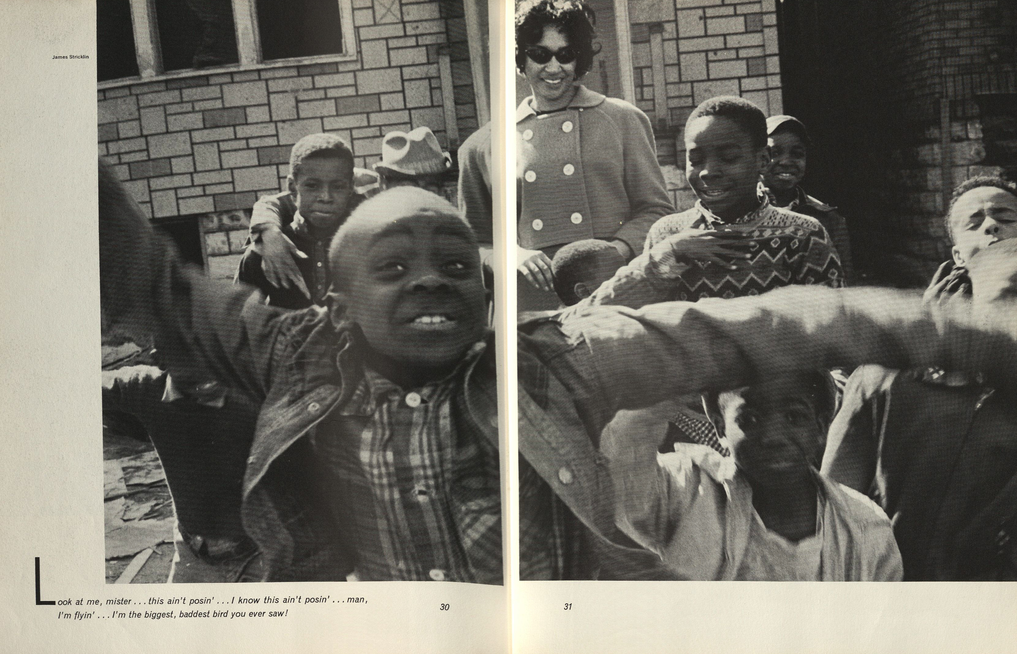 Black-and-white photograph spread across two pages showing a lively group of children and one adult outside a stone-faced building. In the foreground, a young boy leans toward the camera with arms stretched wide, smiling and mid-motion, occupying the center of the composition. Behind him, other children grin and gesture animatedly. At the back, a woman in a buttoned coat and sunglasses stands watching with a smile. The setting appears residential, with brick and stone textures on the surrounding buildings. Printed text at the bottom left reads: "Look at me, mister… this ain’t posin’… I know this ain’t posin’… man, I’m flyin’… I’m the biggest, baddest bird you ever saw!" The name James Stricklin is printed at the top left of the left page.