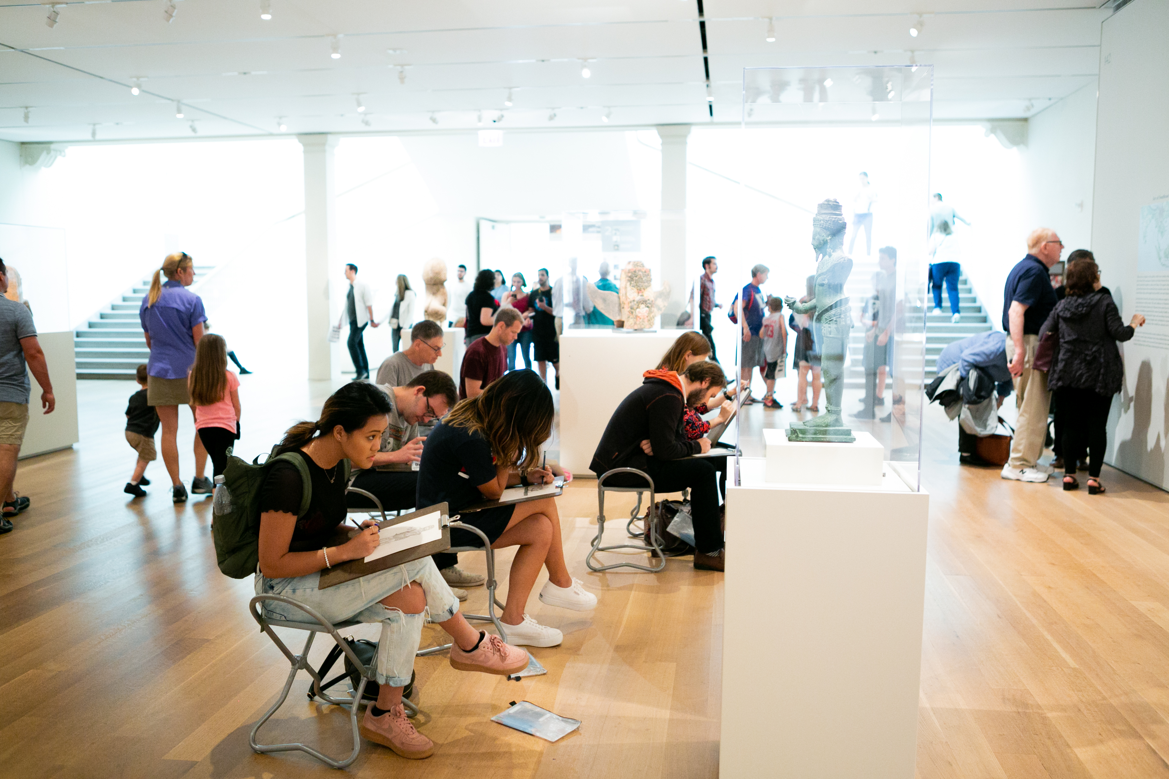 A group of people sit on stools sketching in a white, light-filled art gallery.