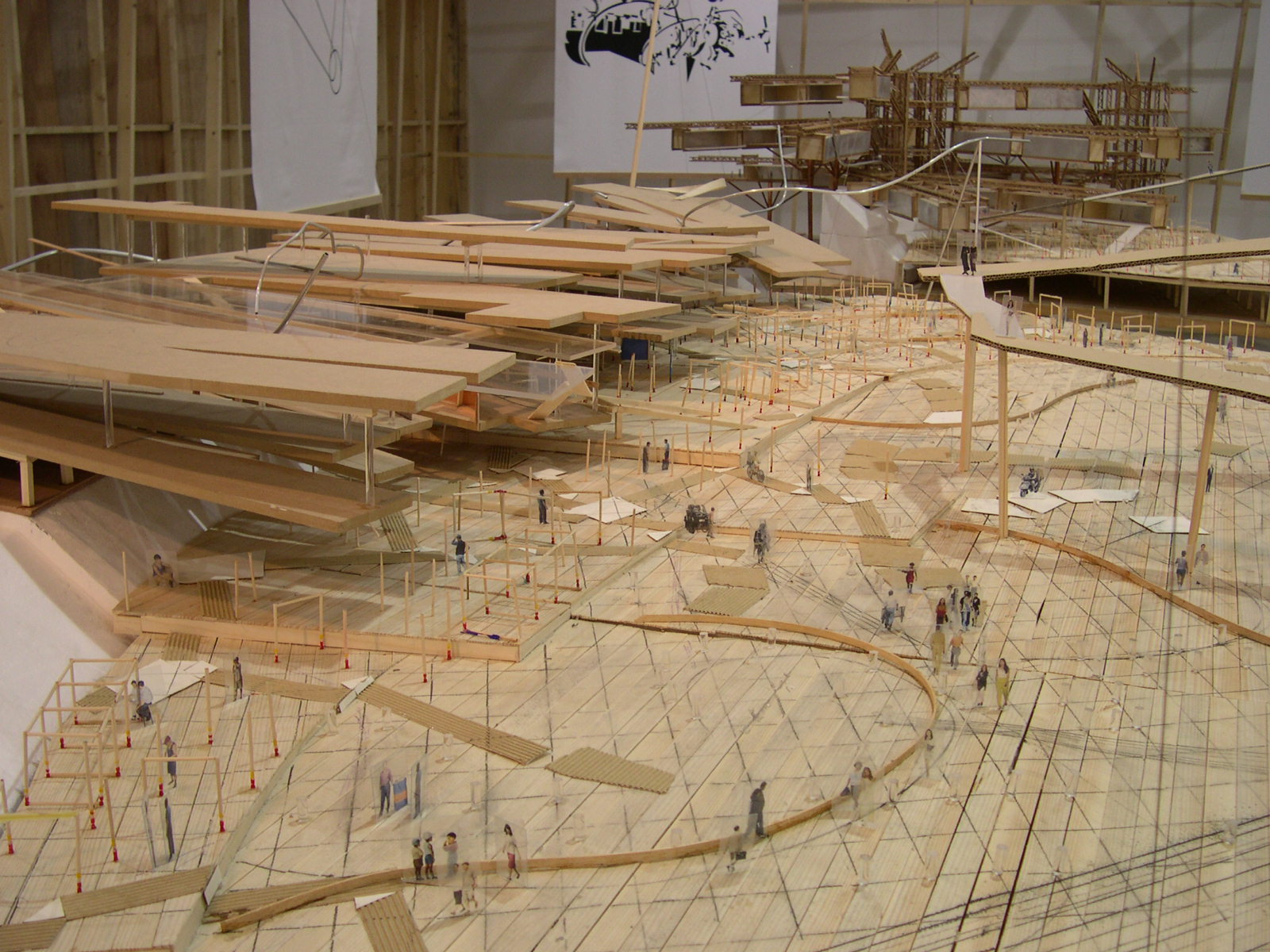 A detail of a model made of cardboard, wood, and string. On the left side, several long, rectangular sheets of wood are supported in the air with metal wire and thin columns made of wood. On the ground  numerous architectural-scale figures seem to walk around small structures made of wood and cardboard.