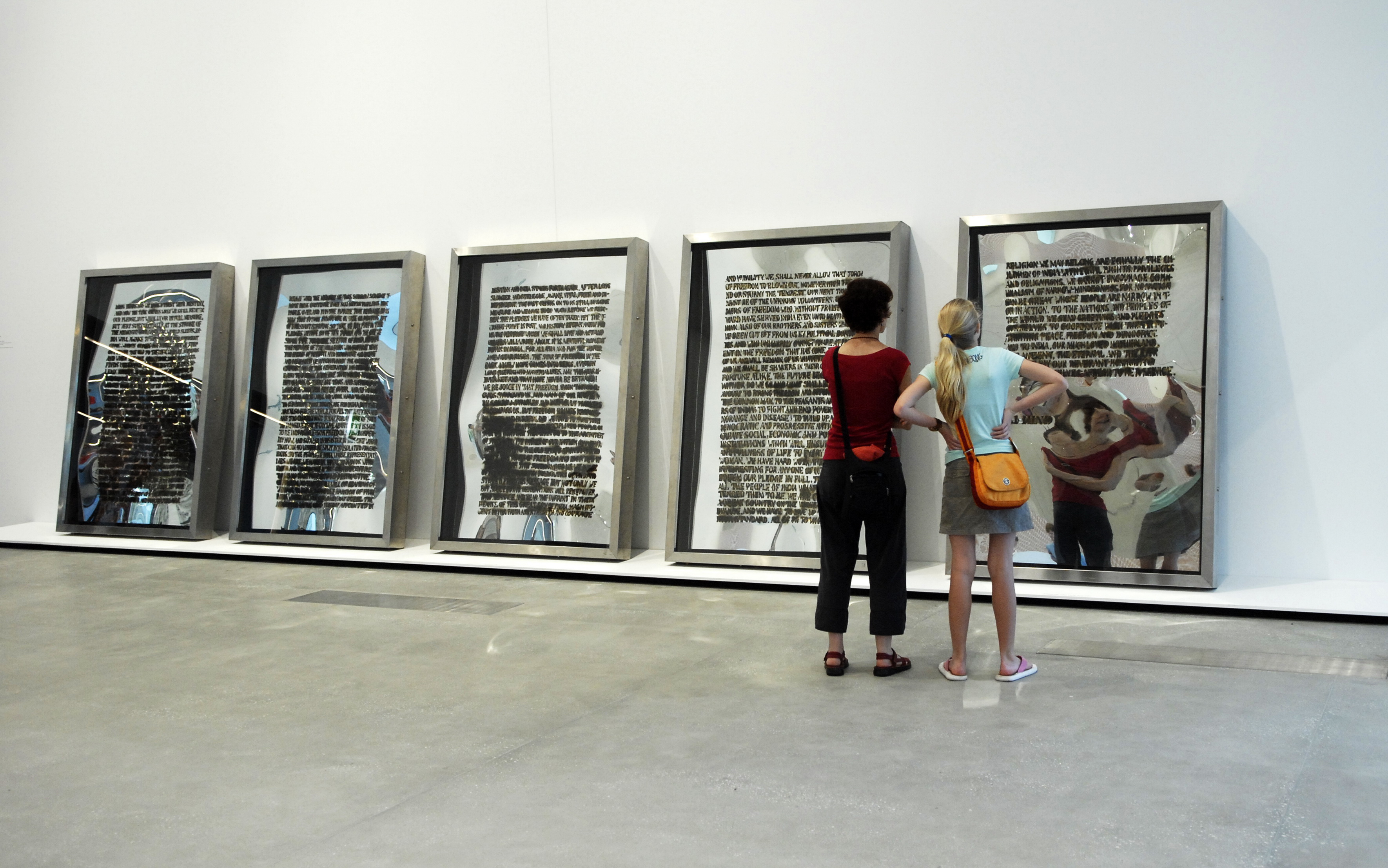 Two viewers stand in front of five large framed mirrors that lean against a white wall. Text in black fills each mirror.