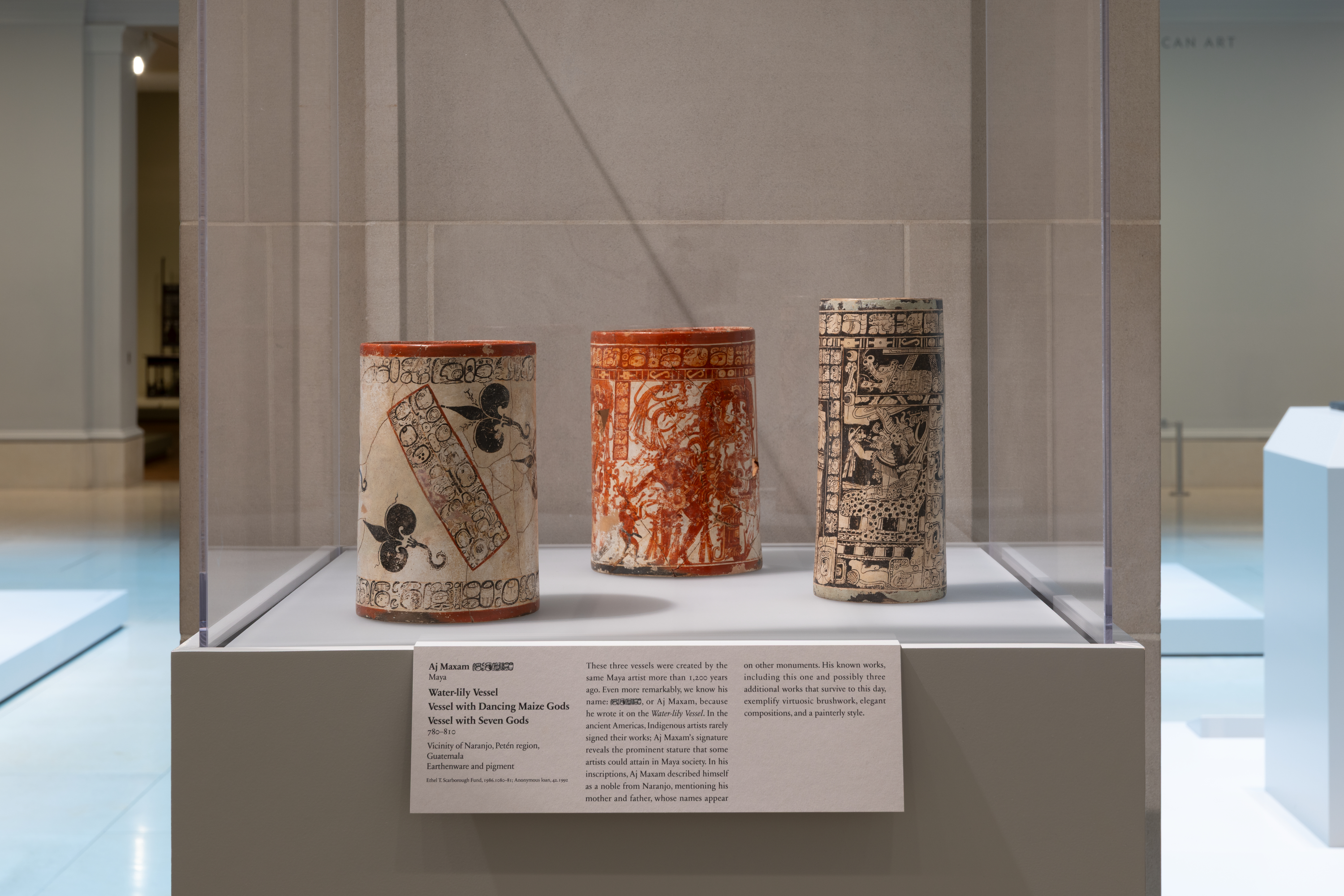 A color photograph shows a museum display case with three tall cylindrical ceramic vessels on a pedestal: at left a cream vessel with a red rim and black motifs, at center an orange-red vessel with dense designs, and at right a cream vessel with black intricate patterns, with a label below.