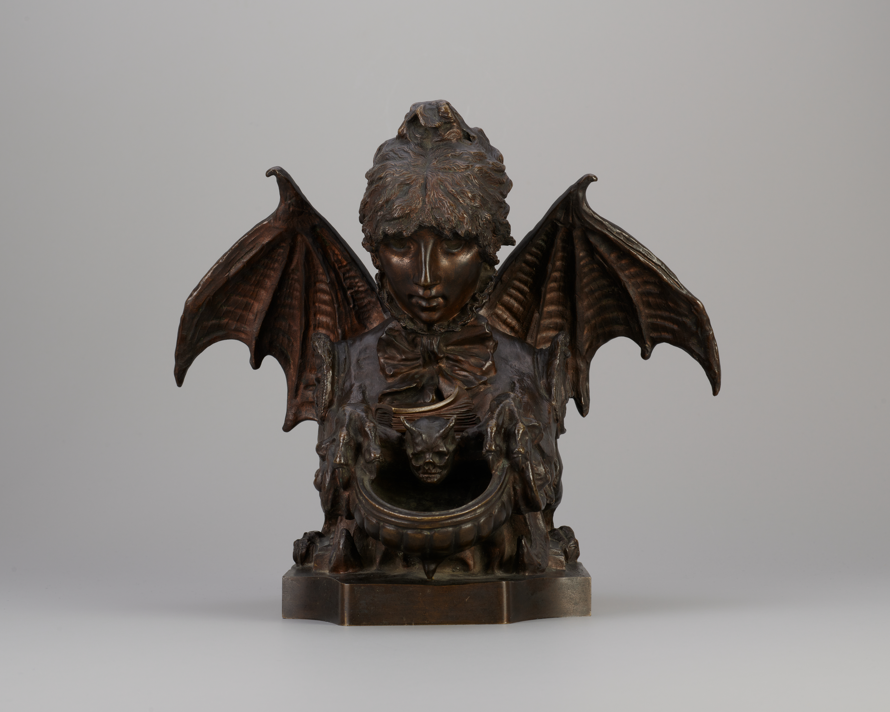 Front view of a bronze-colored metal sculpture of a creature with the head of a curly-haired young woman, extended bat’s wings, and taloned claws. The figure looks down at a book in its lap.