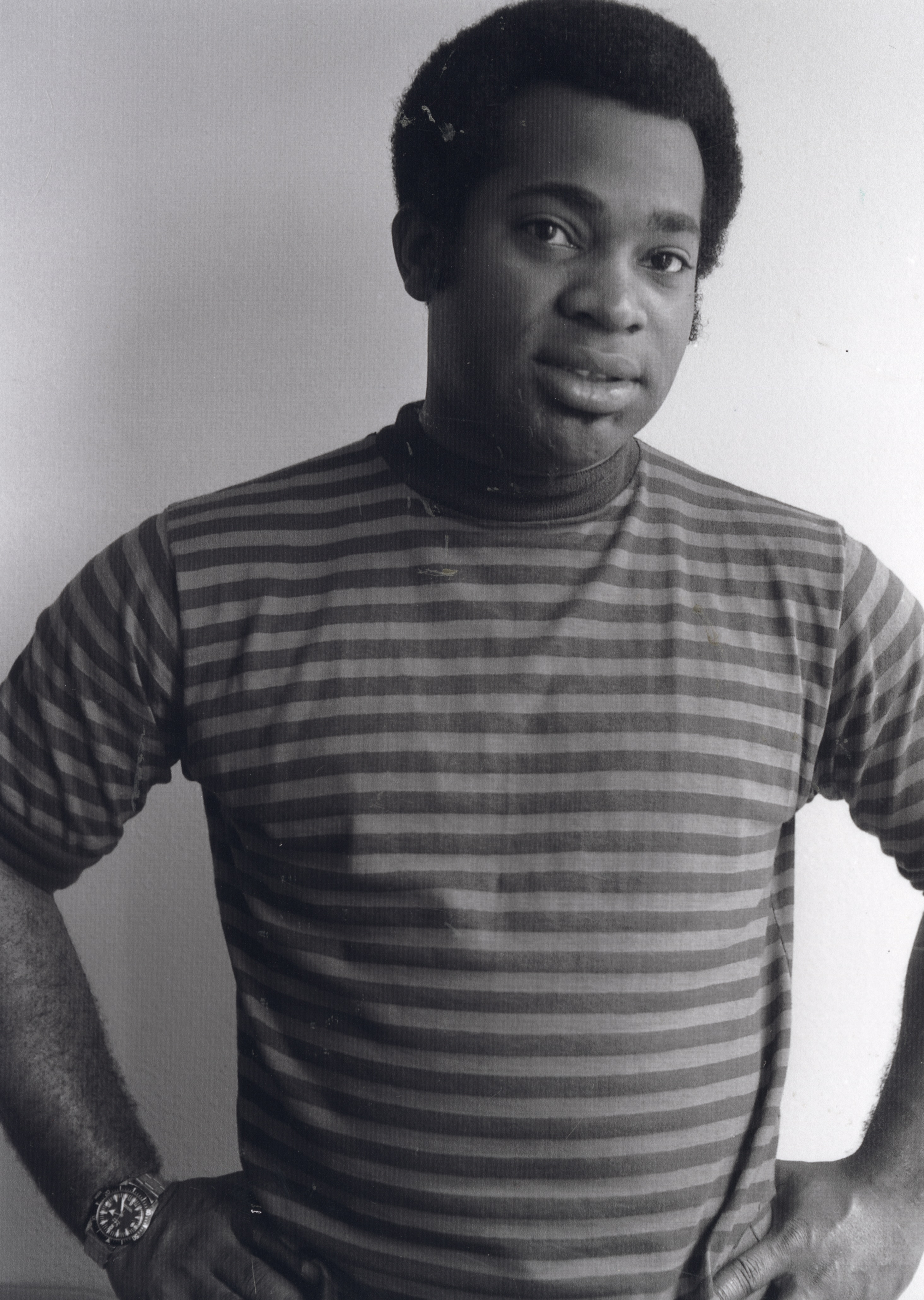 Photograph of a man standing in front of a plain light-colored wall with his hands on his hips. He wears a striped short-sleeve shirt layered over a dark turtleneck and a wristwatch on his left wrist. He has a short afro hairstyle and a calm, direct expression, with his head slightly tilted and eyes looking slightly past the camera.