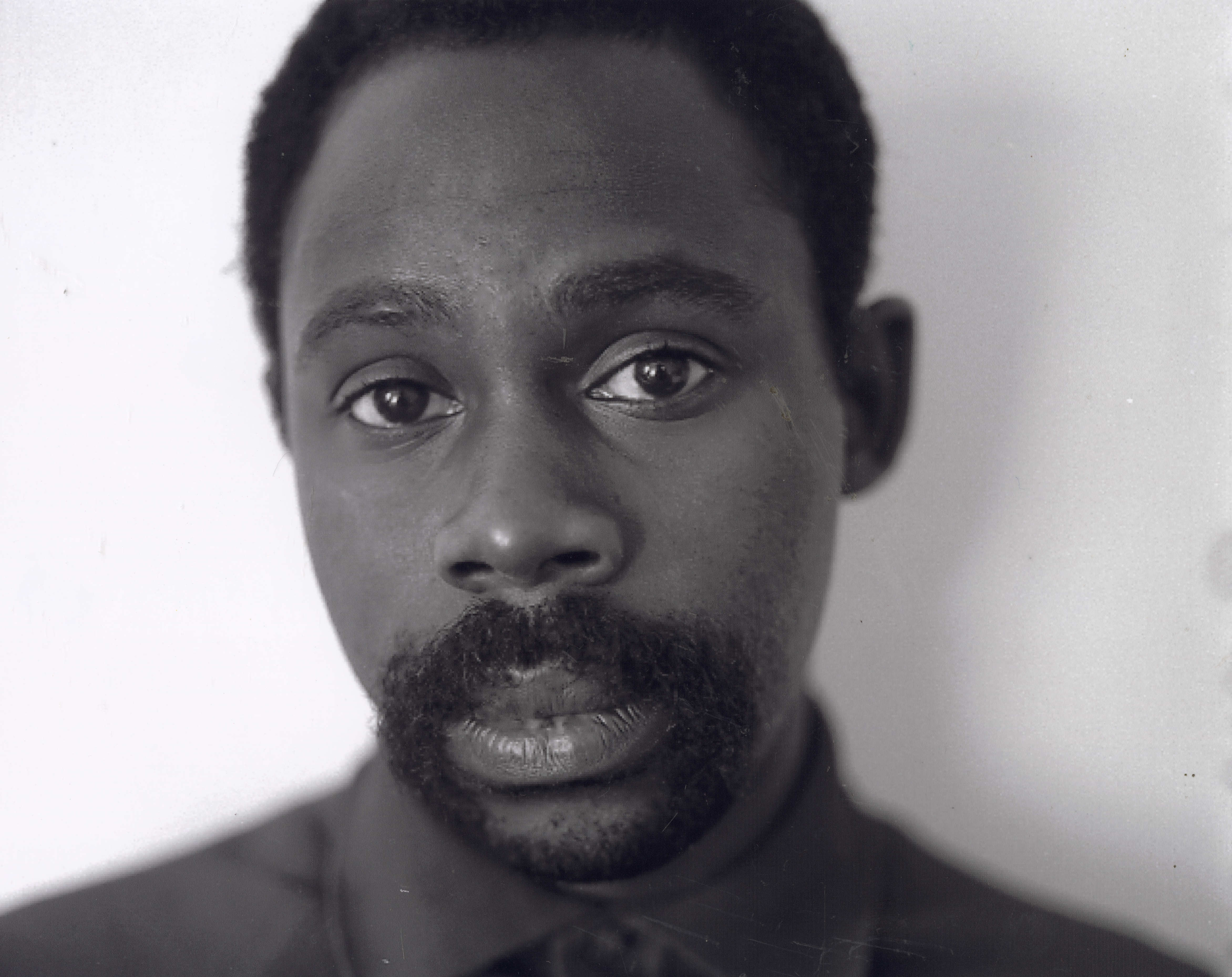 Close-up portrait of a man looking directly into the camera with a serious expression. He has short hair, a full mustache and beard, and wears a buttoned-up dark shirt. The background is a plain light surface, softly lit to emphasize his facial features and direct gaze.