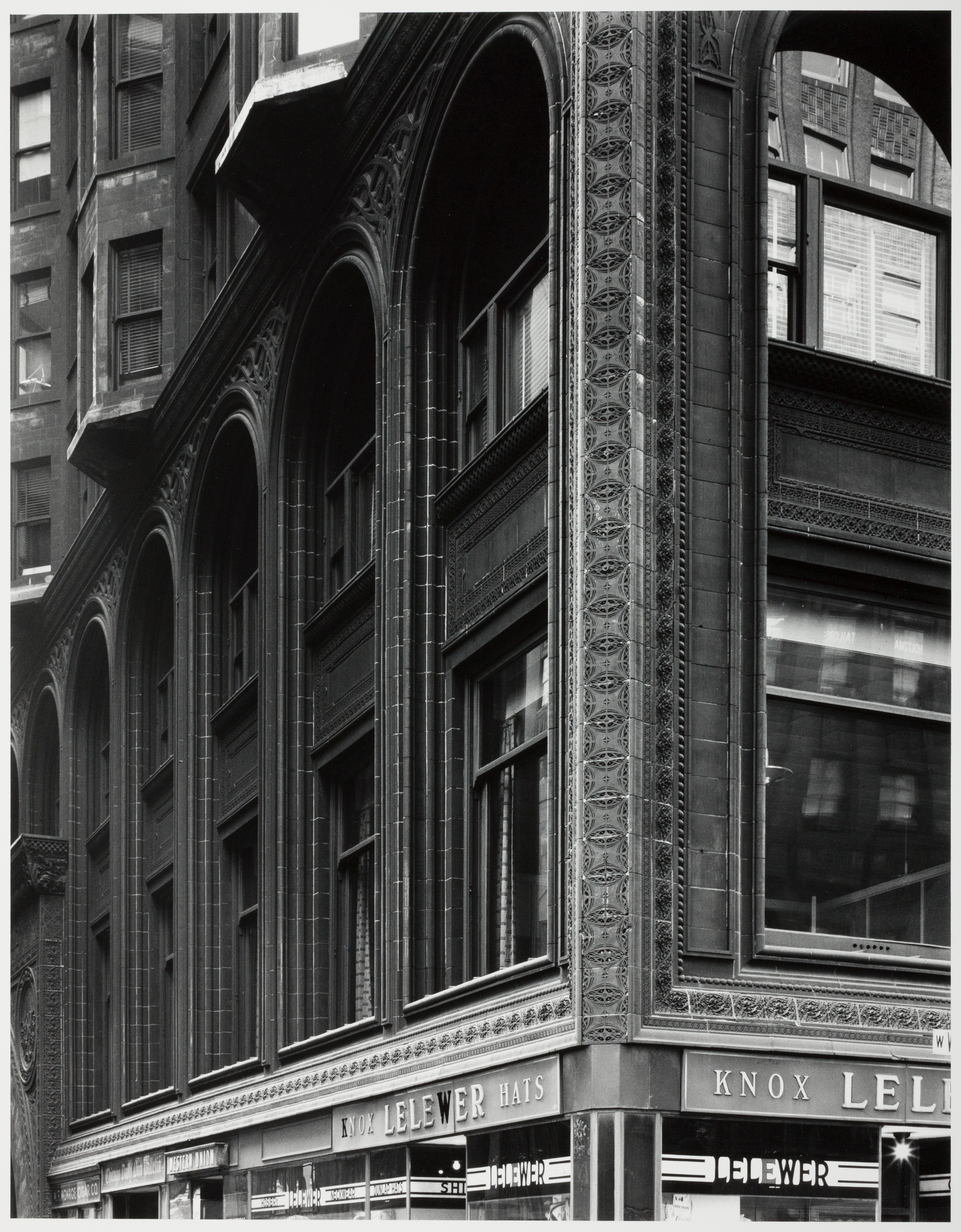Black-and-white photograph of a commercial building facade with arched windows and ornamental detailing. The structure features tall, narrow windows framed by dark tile and decorative trim. The corner is marked by a vertical strip of intricate relief patterns running from the street level to the upper stories. The storefront at street level displays signs reading "KNOX LELEWER HATS" and "LELEWER," with window displays visible below the signage. The photo captures the architectural elegance and texture of a historic urban retail building, highlighting both craftsmanship and commercial identity.