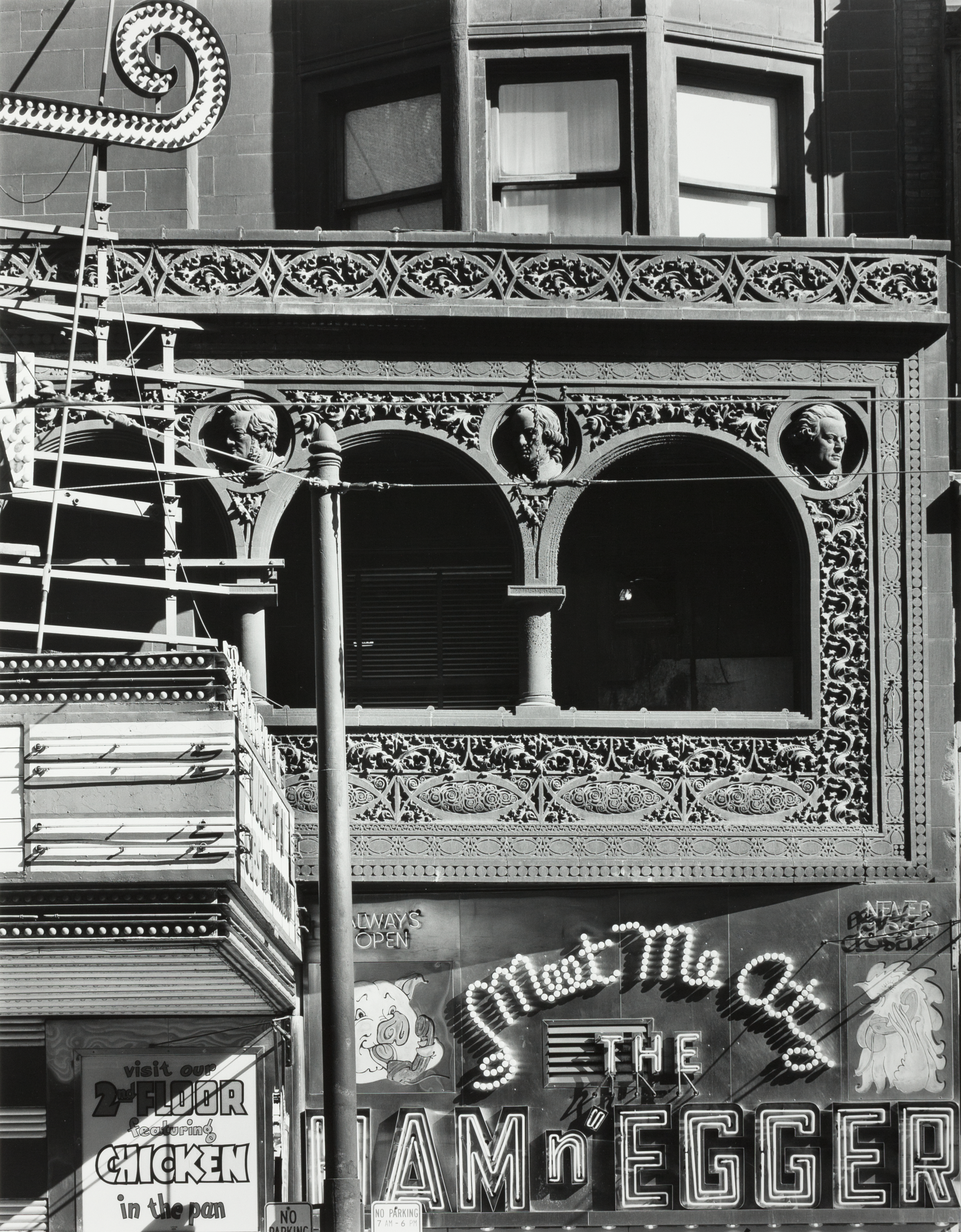 Black-and-white photograph of an ornate building facade featuring carved architectural details and commercial signage. A richly decorated second-story bay is adorned with intricate floral reliefs and three sculpted busts framed by arches. Below, a diner or restaurant sign reads "Meet Me at THE HAM n' EGGER" in bold lettering with stylized neon lights. To the left, part of a vertical marquee and a street pole partially obscure the view. A smaller sign advertises “CHICKEN in the pan” on the second floor. The photograph captures a blend of historic architecture and mid-20th-century street-level commercial culture.