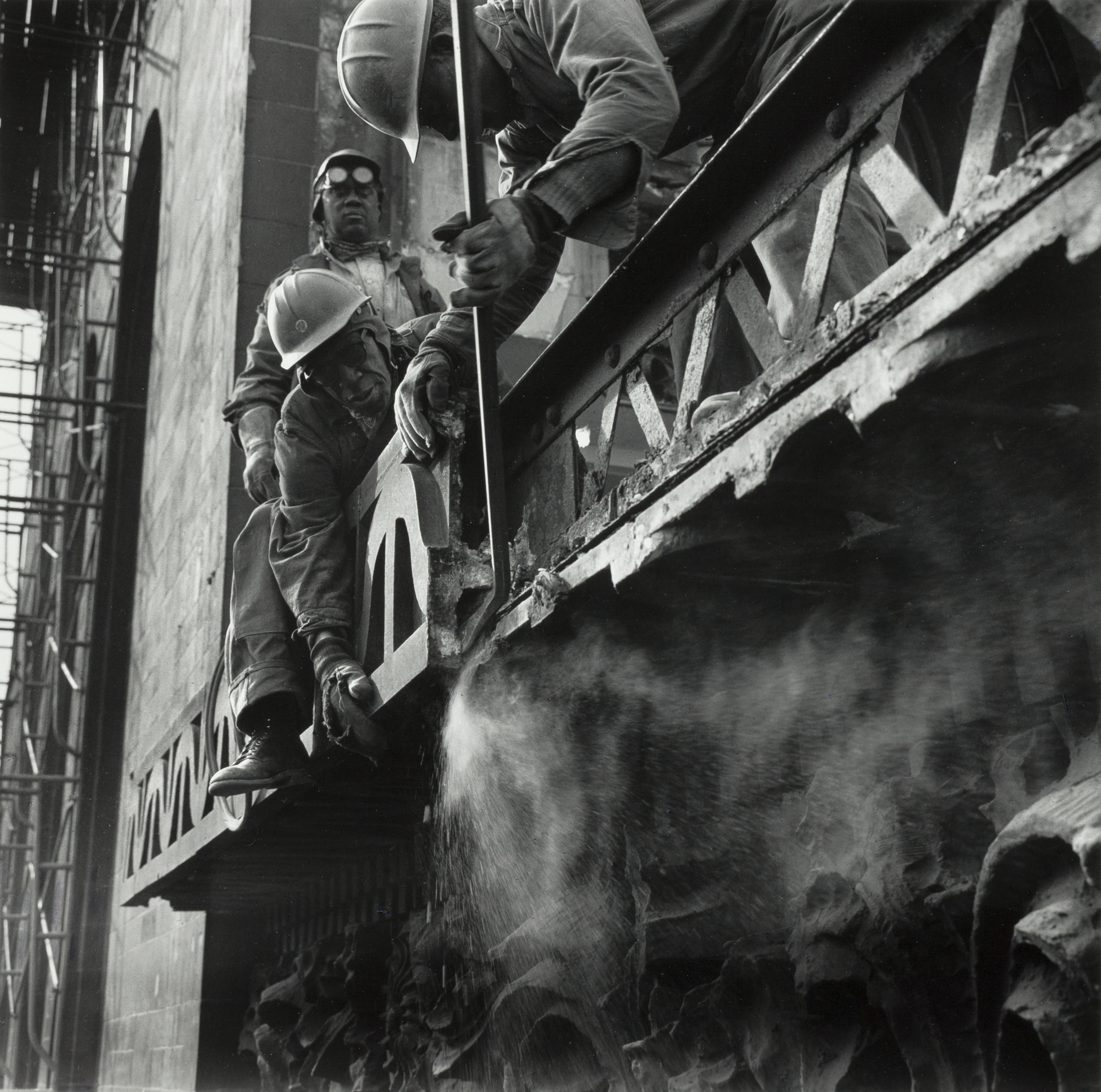 Three construction workers in hard hats and protective gear work on a high ledge of a stone building. One man leans forward using a metal rod to pry part of the decorative structure, while another crouches beside him, and a third stands behind them, facing the camera with goggles and a respirator. A cloud of dust or debris sprays from the ledge as they work. Scaffolding lines the building in the background, emphasizing the scale and height of the restoration or demolition effort.