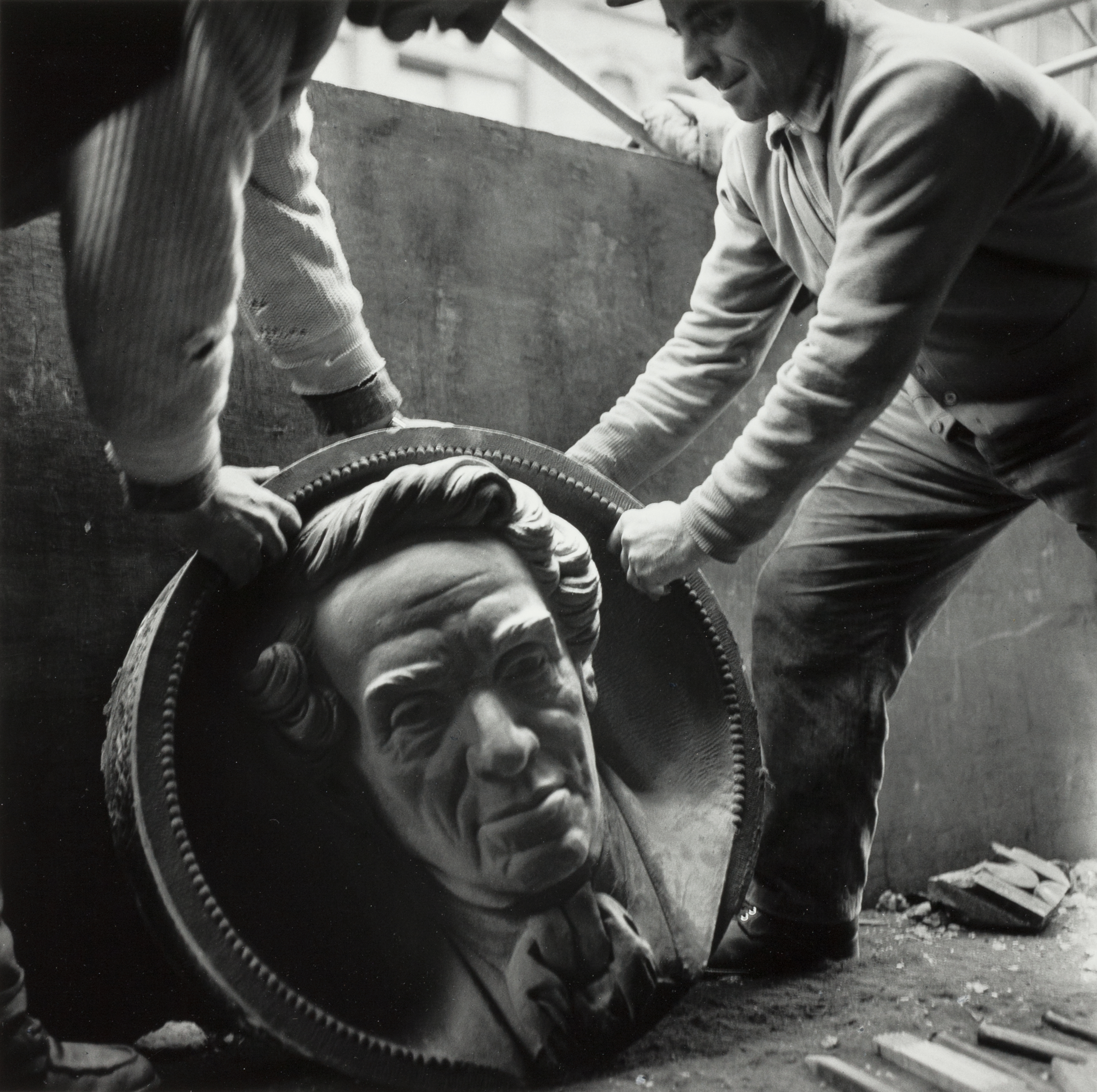 Two men wearing work clothes carefully lift and position a large, round sculpted relief of a man's face framed by a beaded circular border. The portrait features stylized hair and a cravat, suggesting a historical figure. The setting appears to be a workshop or construction site, with debris and tools scattered on the ground. The composition emphasizes the careful handling of the detailed artwork.