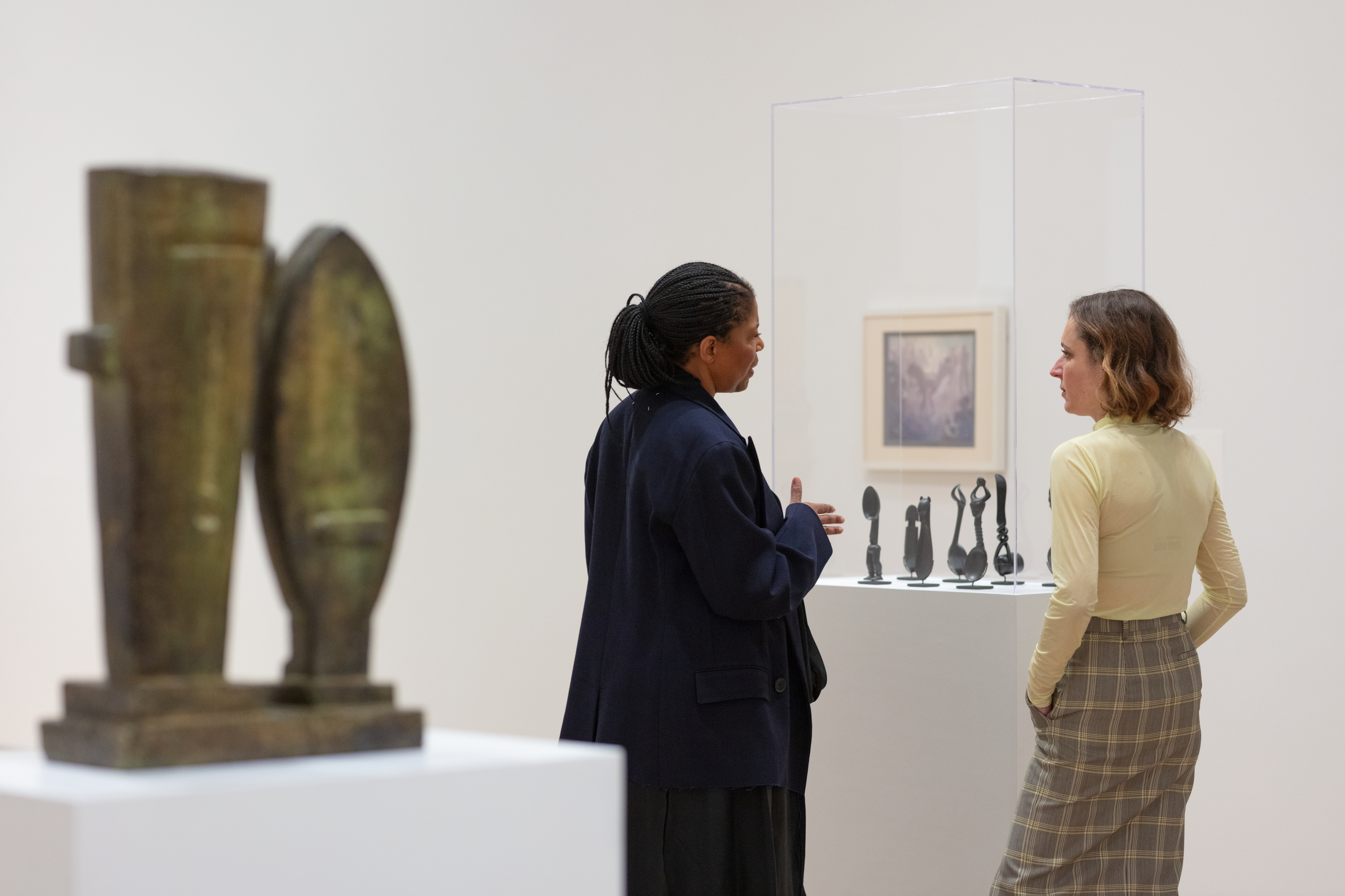 Two women, Simone Leigh—a Black woman with braids in a structured navy sportcoat—and Paulina Pochoba—a light-skinned woman with short, wavy highlights and a yellow turtleneck—stand together in a white gallery space, their backs turned mostly to the viewer, their faces in profile. Leigh gestures toward a grouping of small metal objects in a virtine. To her left, close up and blurry, is is a geometric sculpture in bronze tones.