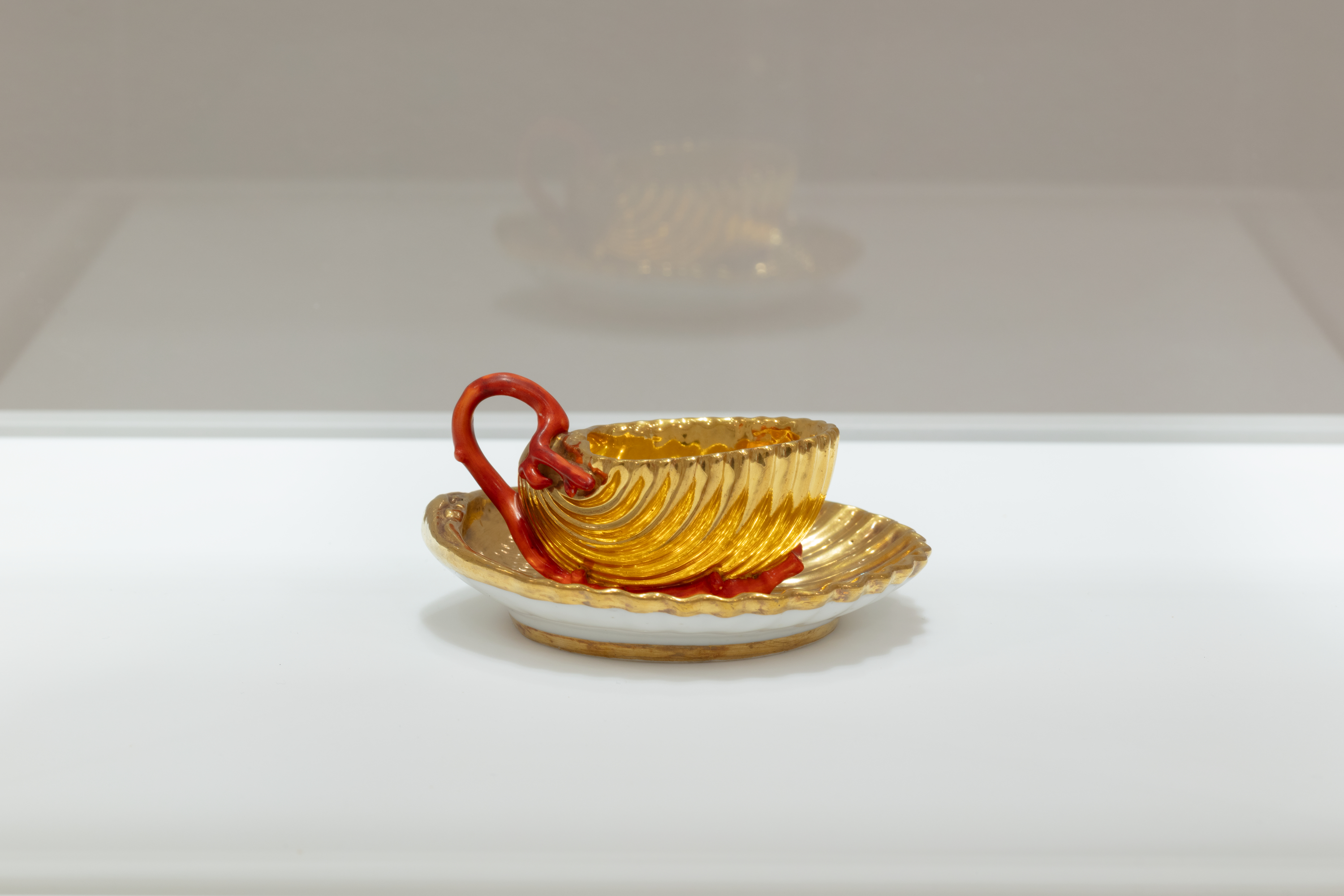 A gilded teacup set on a golden saucer, both with a scalloped surface. The cup sports a delicate, organically shaped red-orange handle.