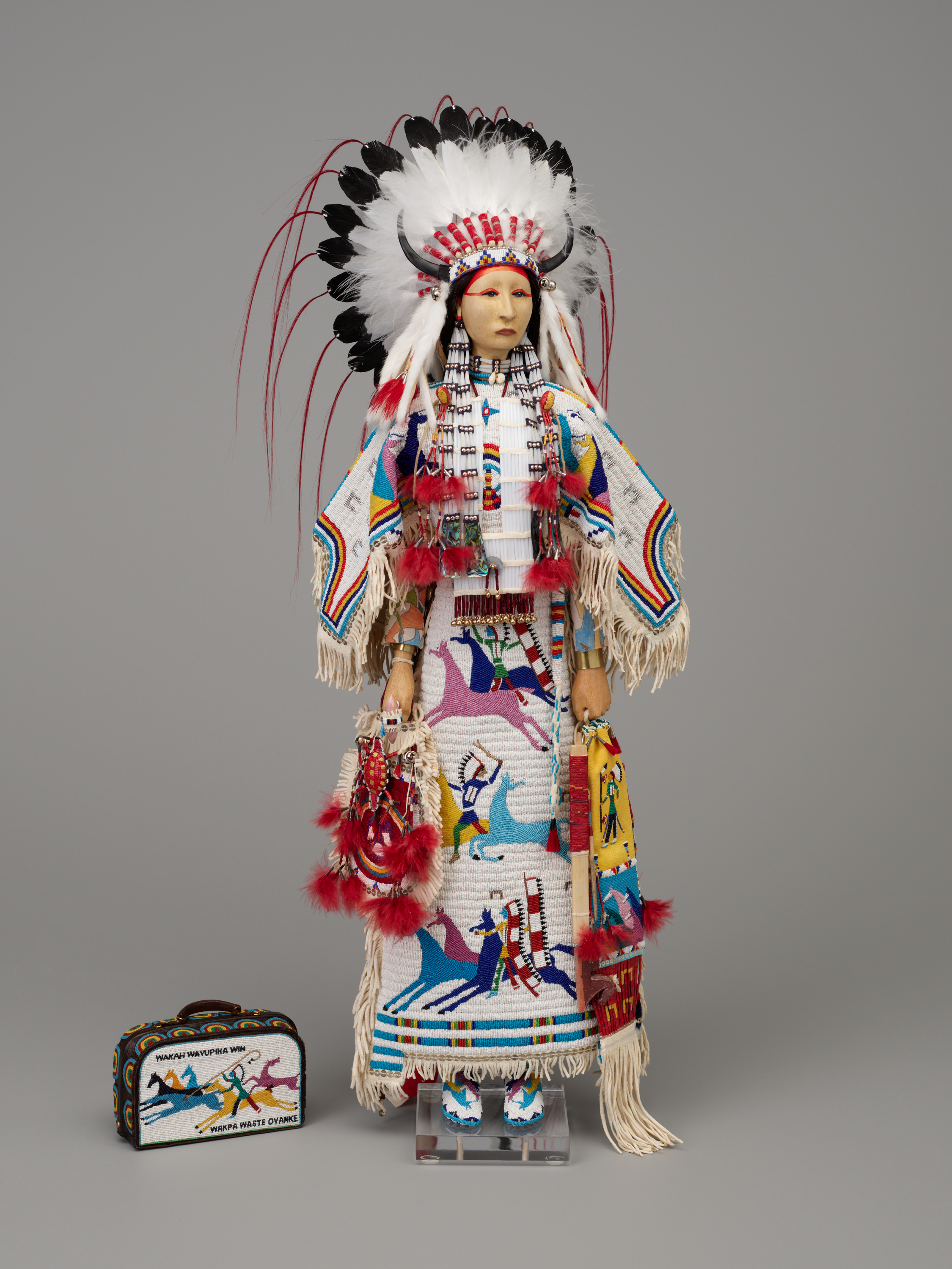 A figure of a woman dressed in a feathered war bonnet with horns and an elaborately beaded robe featuring images of animals in pink, blue, yellow, red, and black. A small case with an animal design on it rests next to her.