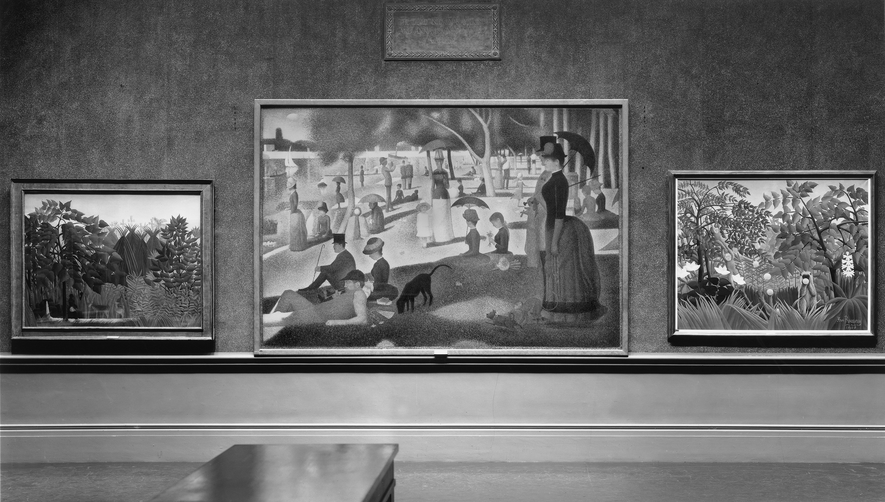 Black-and-white photograph of three paintings hung in a dark gallery, the centermost being Seurat's "La Grande Jatte" in a thin, light frame.