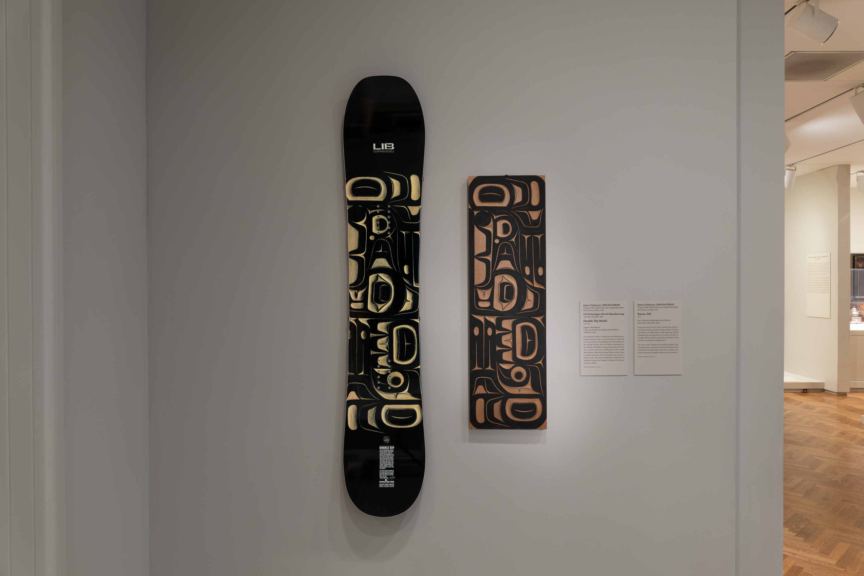 A color photograph in a gallery shows, at left, a black snowboard with tan abstract shapes mounted on a gray wall; at center-right, a narrow brown panel with matching tan-and-black pattern hangs beside two white wall labels; gallery floor and lights appear at far right.
