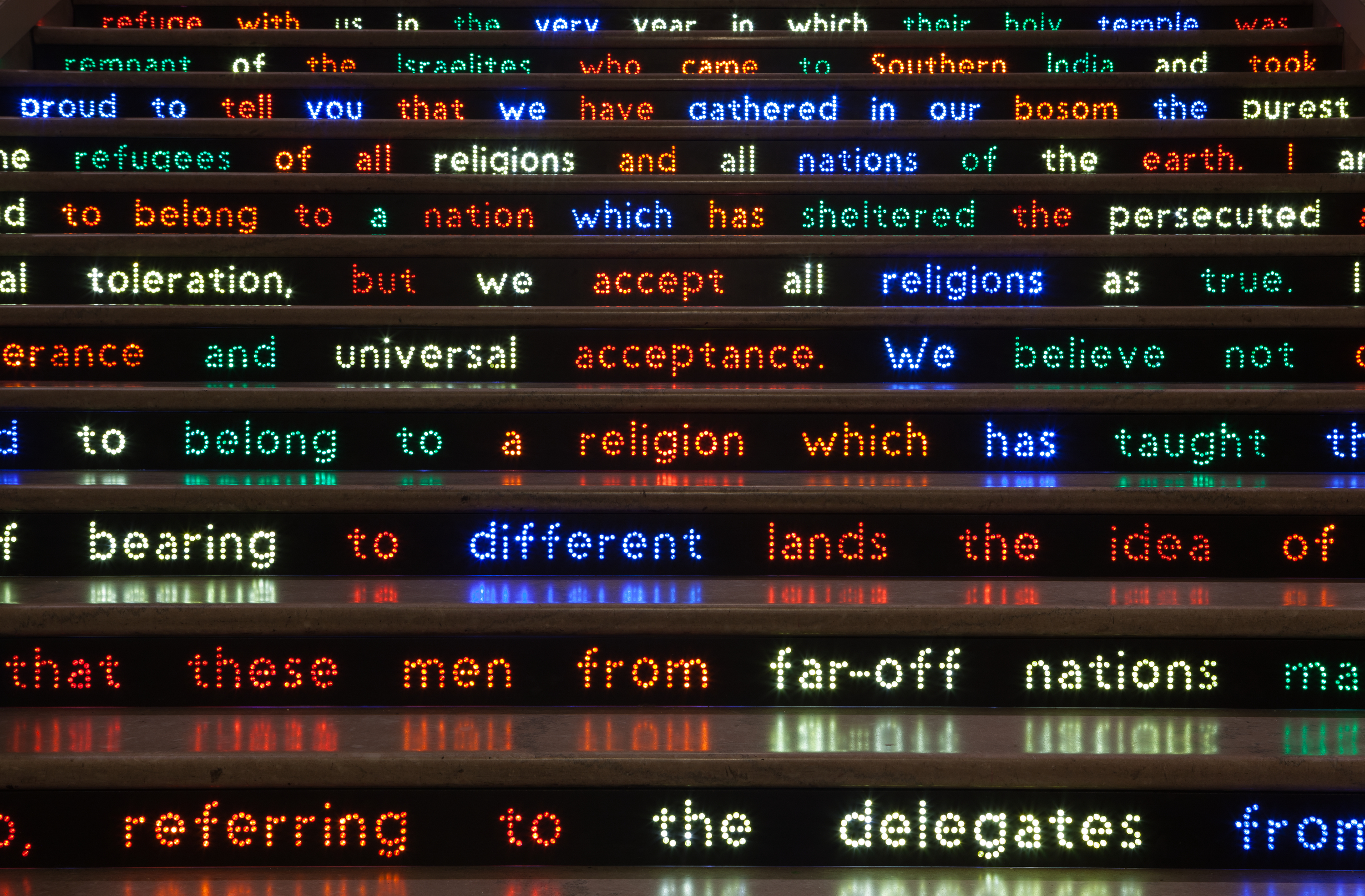 A close-up view of white marble stairs showing the risers, on which words in alternating red, yellow, orange, green, and blue appear against a black background.