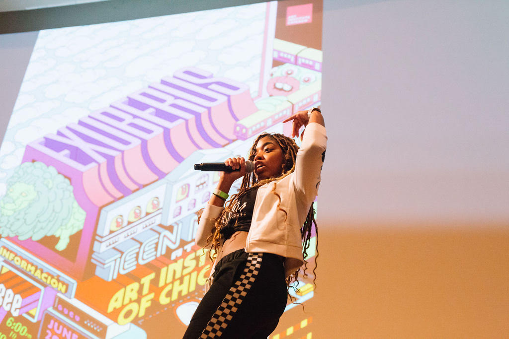Photograph of a medium-skinned teenager in long light-brown braids performing into a microphone, the word "Exuberus" projected behind her in a colorful digital illustration.