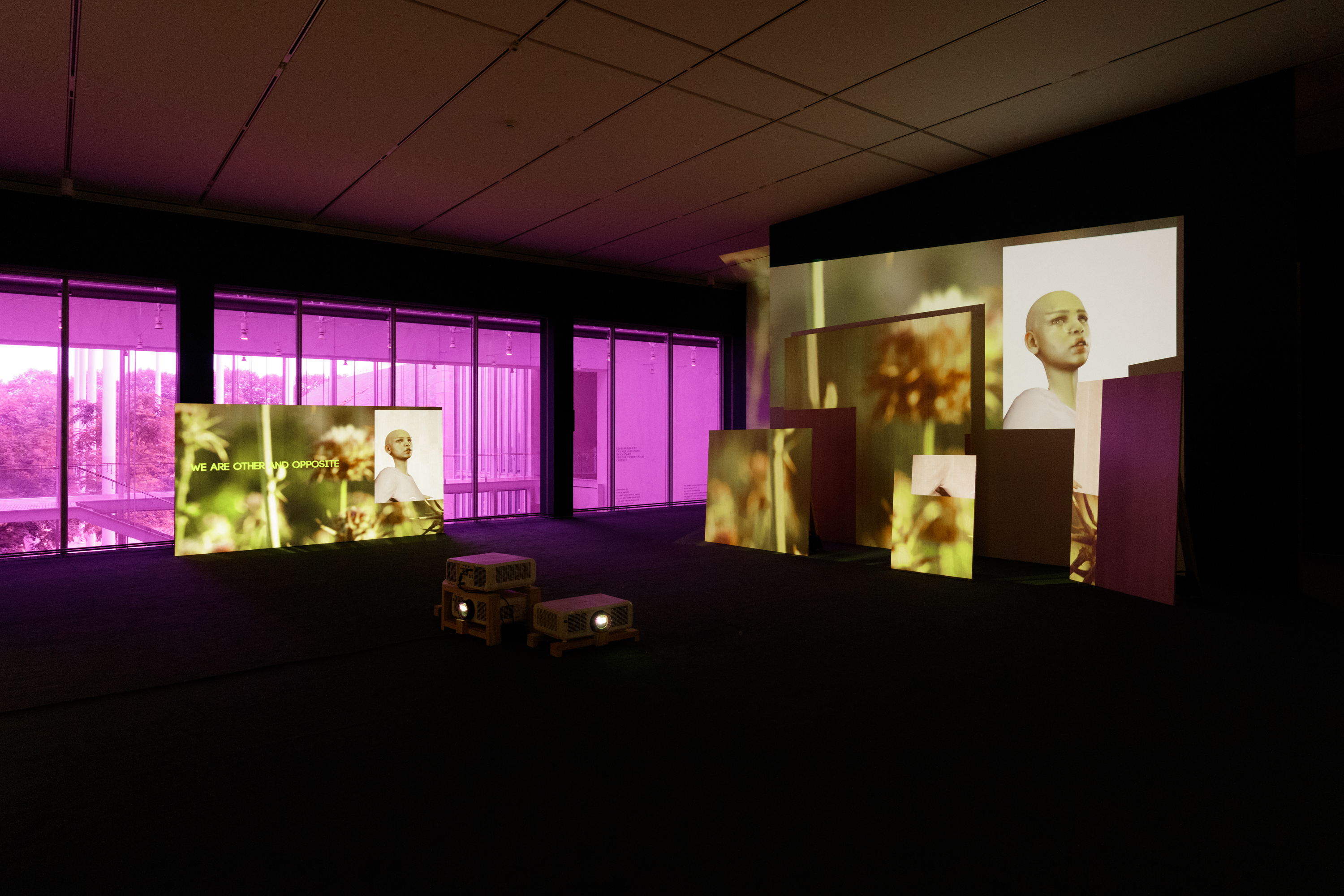 A dark room with a few projectors in the center includes several different sized screen, each showing a different image. A couple include text as well as a digitized image of woman.