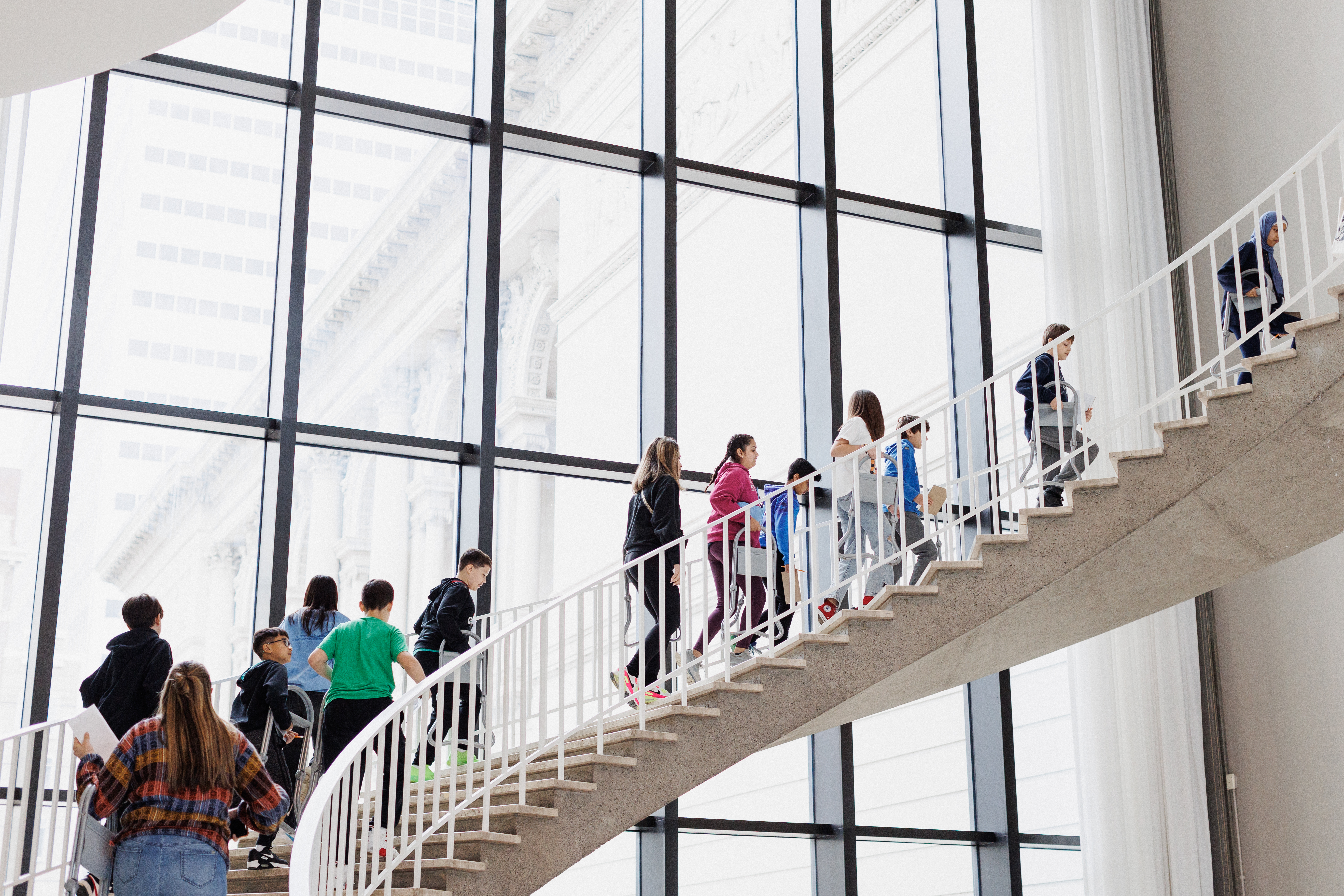 A group of older children walk up a large spiral staircase carrying folding stools. Behind them, floor-to-ceiling windows reveal the pale outline of a stone building with columns.