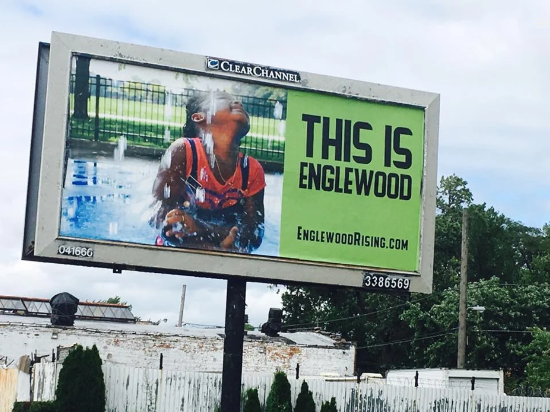 Color photograph of a roadside billboard with a split design. On the left side is a photograph of a smiling child in a red outfit standing under spraying water, possibly in a splash pad or park, with water droplets caught midair. Behind the child is a black metal fence and green lawn. On the right side, a bright green background features bold black text that reads "THIS IS ENGLEWOOD" and below it "EnglewoodRising.com." The billboard is mounted on a black pole with identifying numbers and a Clear Channel label across the top. Trees, utility poles, and rooftops are visible in the background.