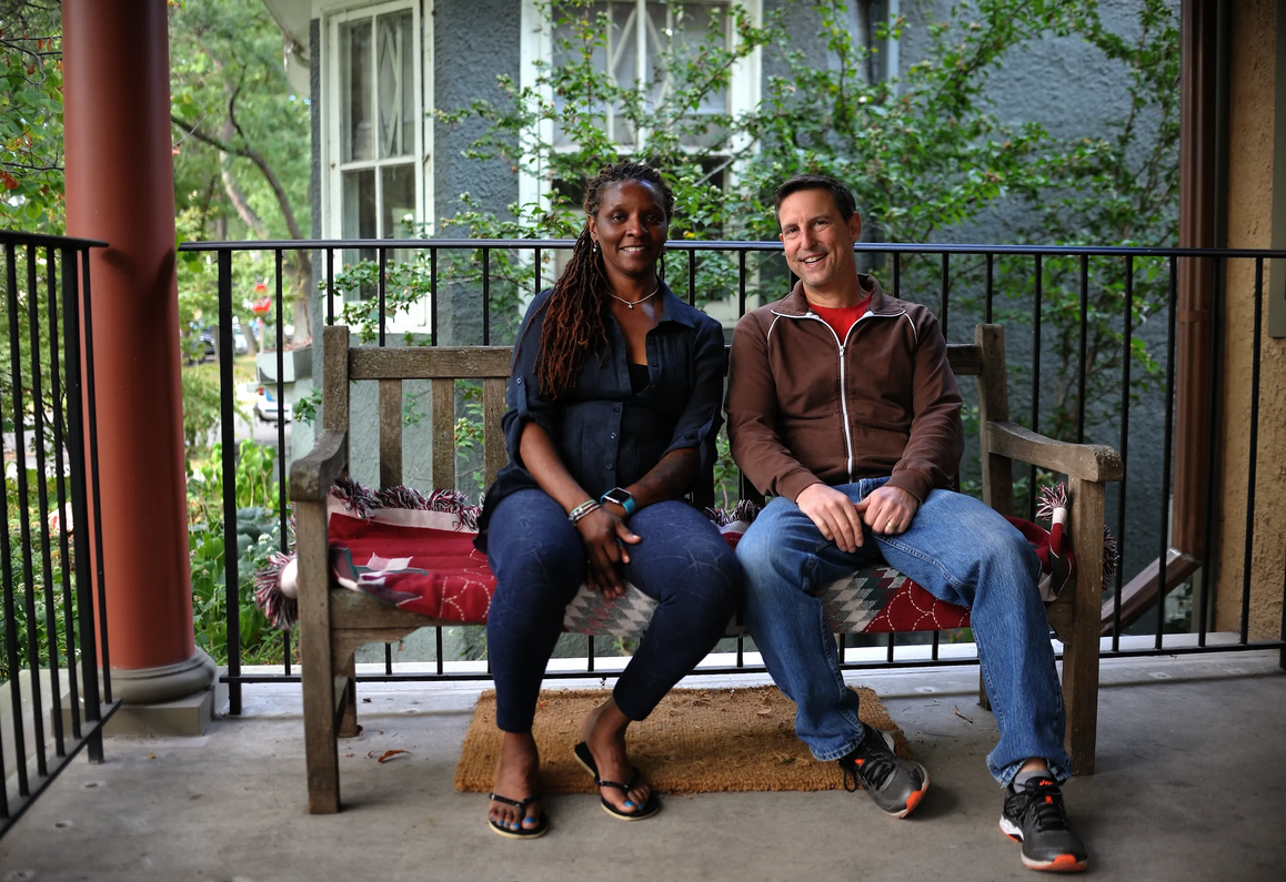 Color photograph of two people sitting on a wooden bench on a porch. The person on the left has long, dark dreadlocks and is wearing a black button-up shirt and dark jeans, smiling with hands resting in their lap. The person on the right has short dark hair and is wearing a brown zip-up hoodie over a red shirt and blue jeans, also smiling with hands folded. The bench has a red and gray patterned blanket draped over the seat. Behind them is a leafy background with trees, a building with textured gray stucco, and a window. A red column and black metal railing frame the porch.