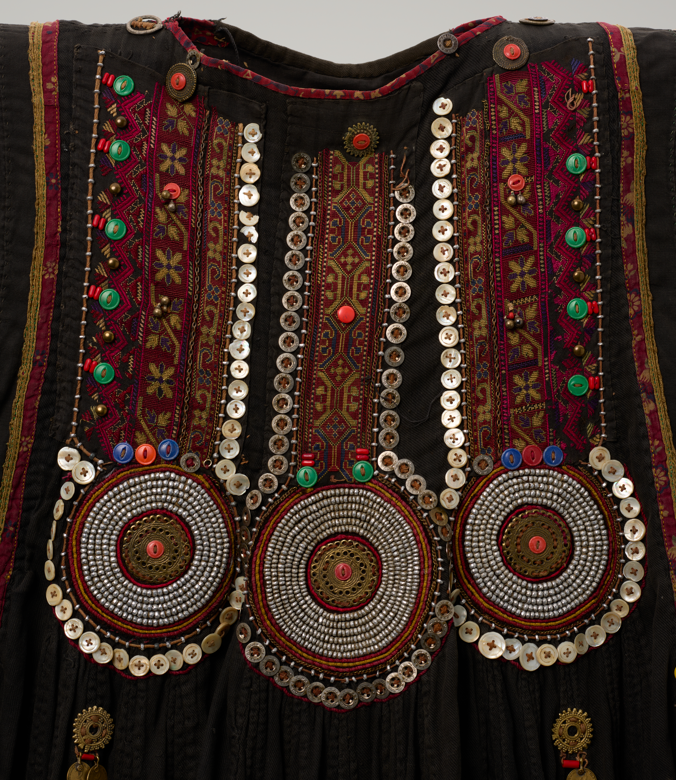 Top edge shows a rounded neckline on a dark garment, trimmed with red and dotted with a few small round buttons. Below it, three vertical bands run down the front, each flanked by rows of white buttons and patterned strips in red, gold, and black, with scattered green, red, and blue buttons along the sides. Across the lower chest, three large round designs sit side by side—concentric rings of silver-colored discs with red and gold centers—each bordered by a ring of white buttons; the middle circle is slightly larger than the two on the sides. The base fabric is black, with narrow red and yellow stripes at the far left and right.