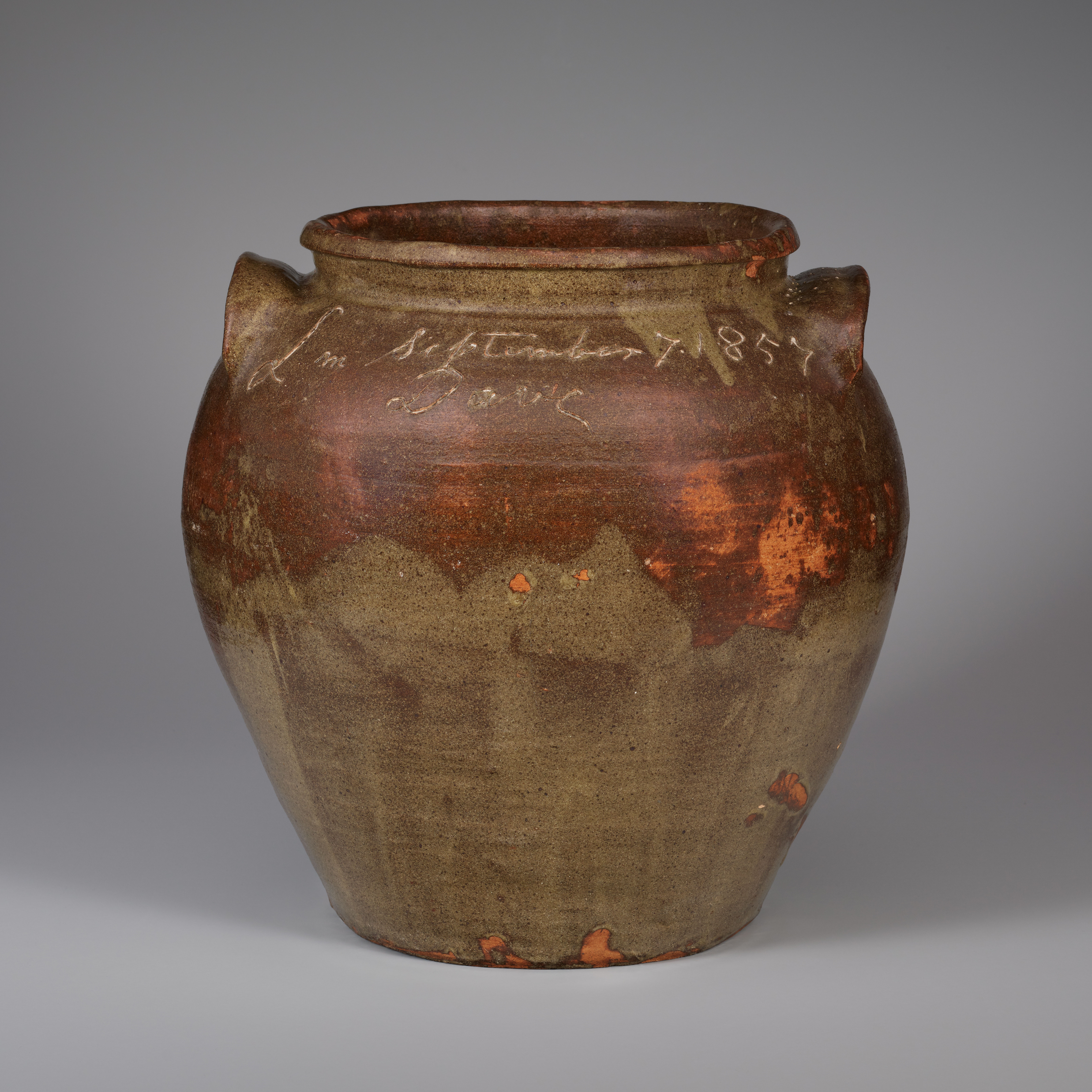 A large ceramic vessel with a wide open mouth and two small handles is inscribed at the top and glazed in brown with green petal shapes overlapping at the bottom and spots of bright orange.