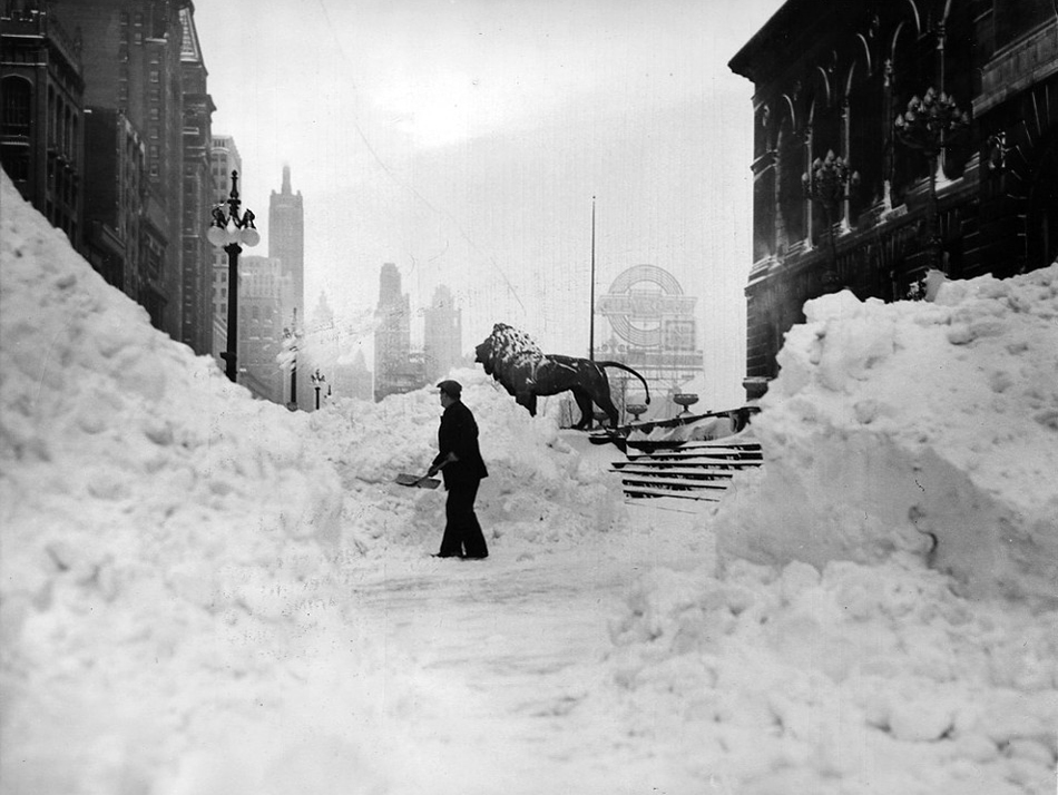 A black-and-white photo in which a person shovels snow in front of the Art Institute steps. The pile is as high as the head of of of the huge lion sculptures in front of the building.