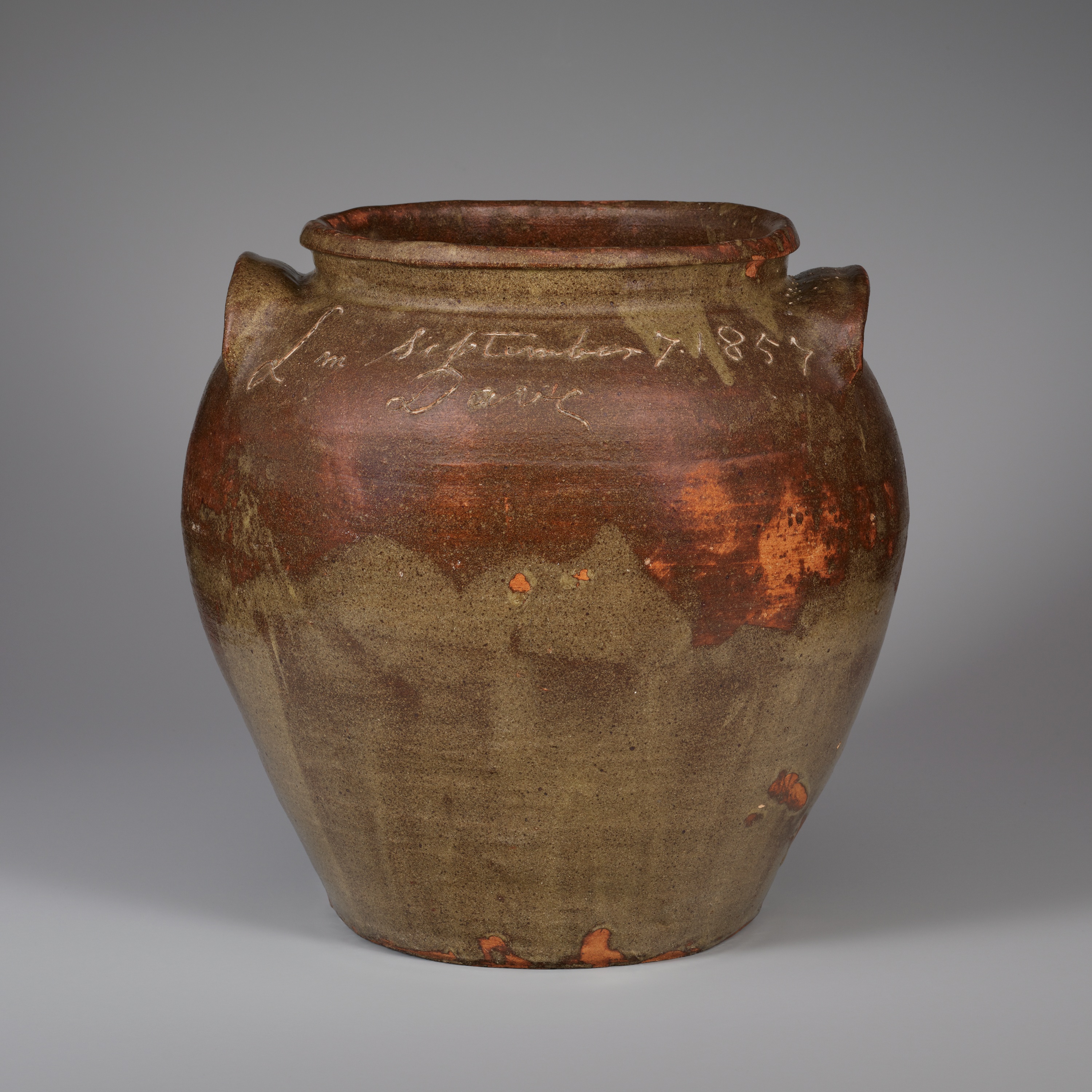A large ceramic vessel with a wide open mouth and two small handles is inscribed at the top and glazed in brown with green petal shapes overlapping at the bottom and spots of bright orange.