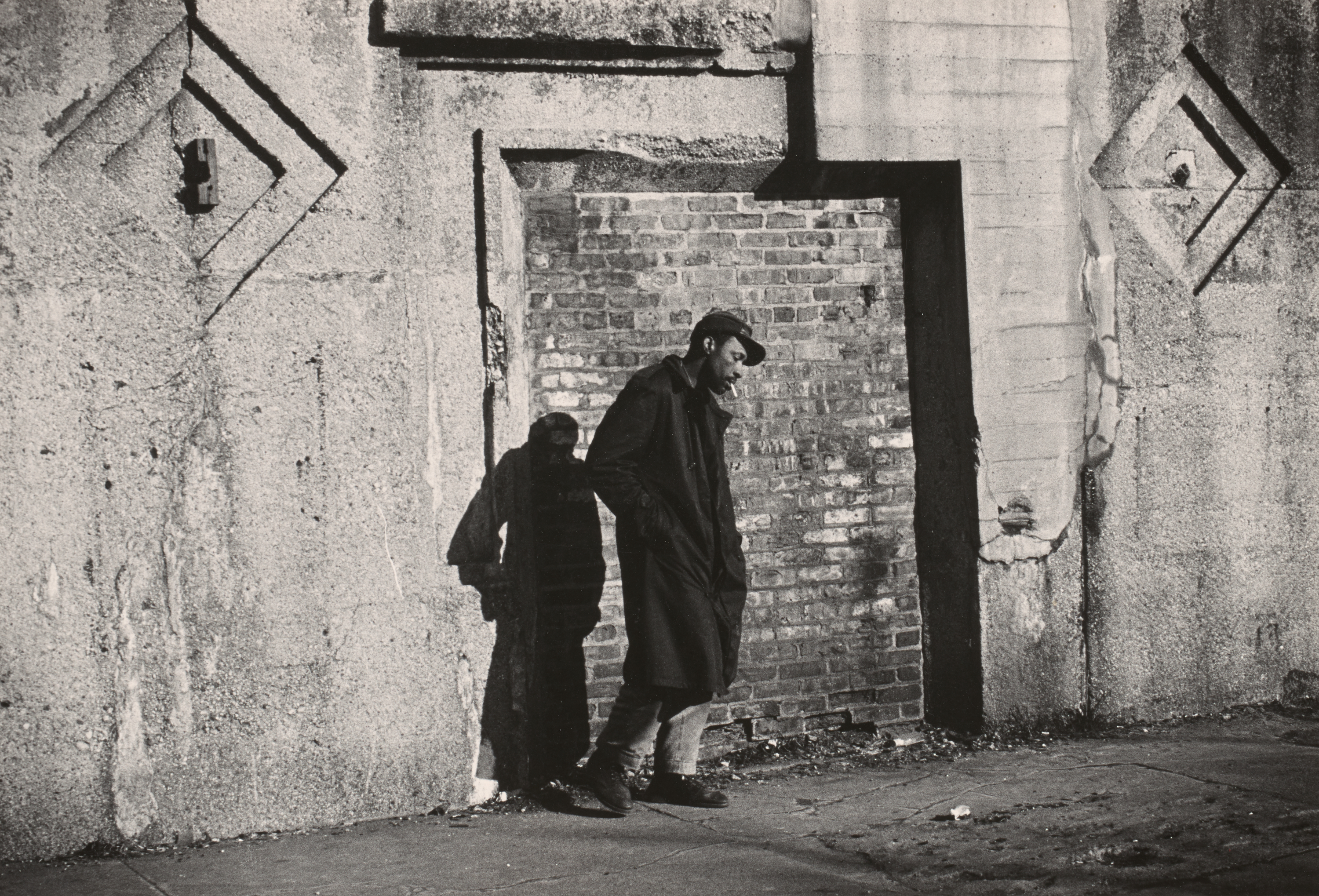 Black-and-white photograph of a man standing alone against a weathered wall made of concrete and brick. He leans back with his head slightly bowed, hands in the pockets of his long dark coat, and a cigarette hanging from his mouth. He wears a cap and rolled-up jeans with boots. The brickwork behind him is partially recessed into the concrete, forming a stark rectangular frame around his figure. The man's shadow is cast on the wall to his left, adding depth and emphasizing his stillness. The setting appears urban and quiet, with strong afternoon light creating dramatic contrast.