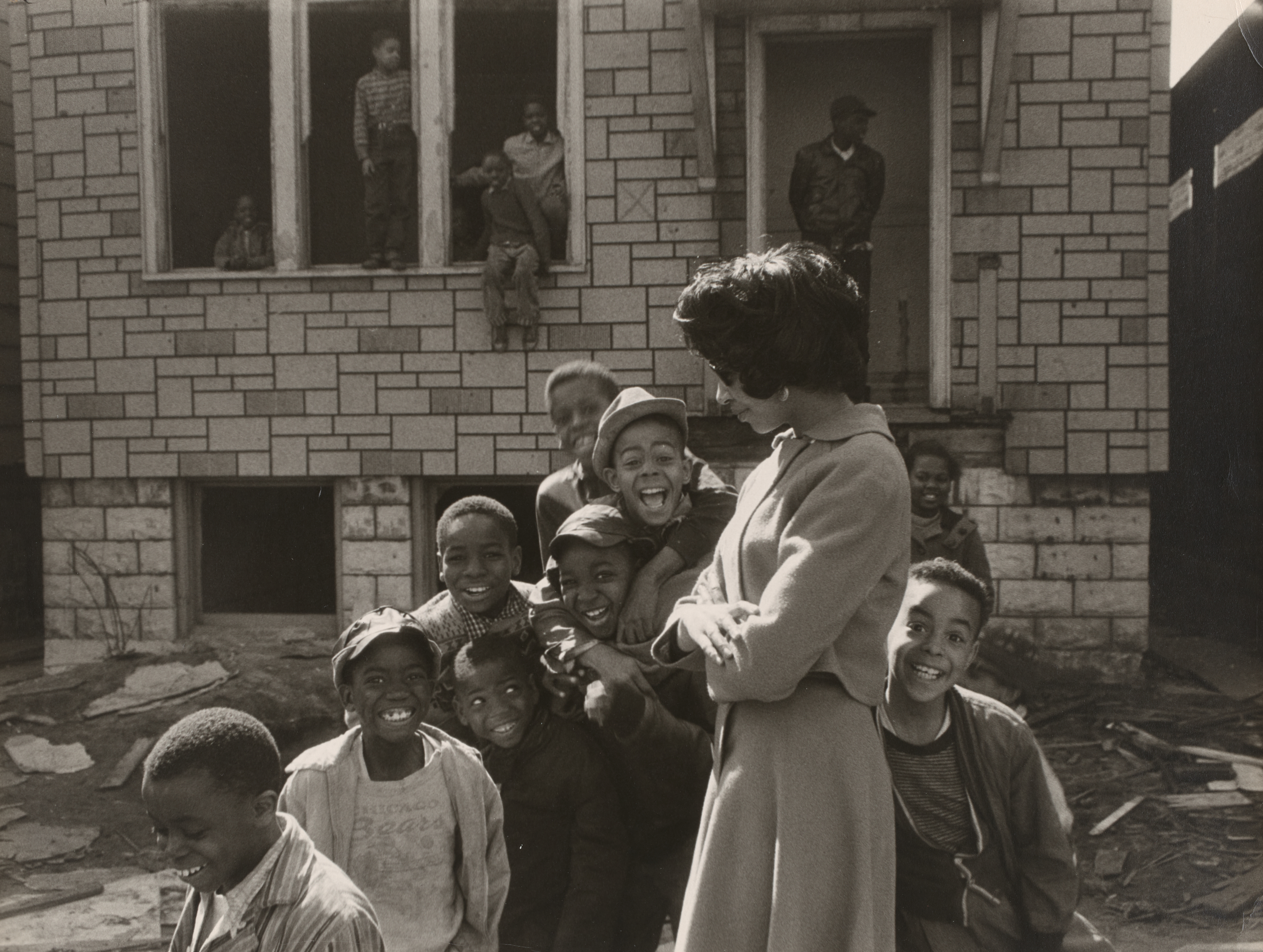 Black-and-white photograph of a group of children gathered around a woman in a coat standing with her arms crossed, smiling and looking down. The children cluster close to her, many grinning or laughing. Behind them is a stone-faced building with missing windows and a broken front step. Several other children are visible in the background, sitting in the window frames or standing on the steps. The ground is littered with debris.