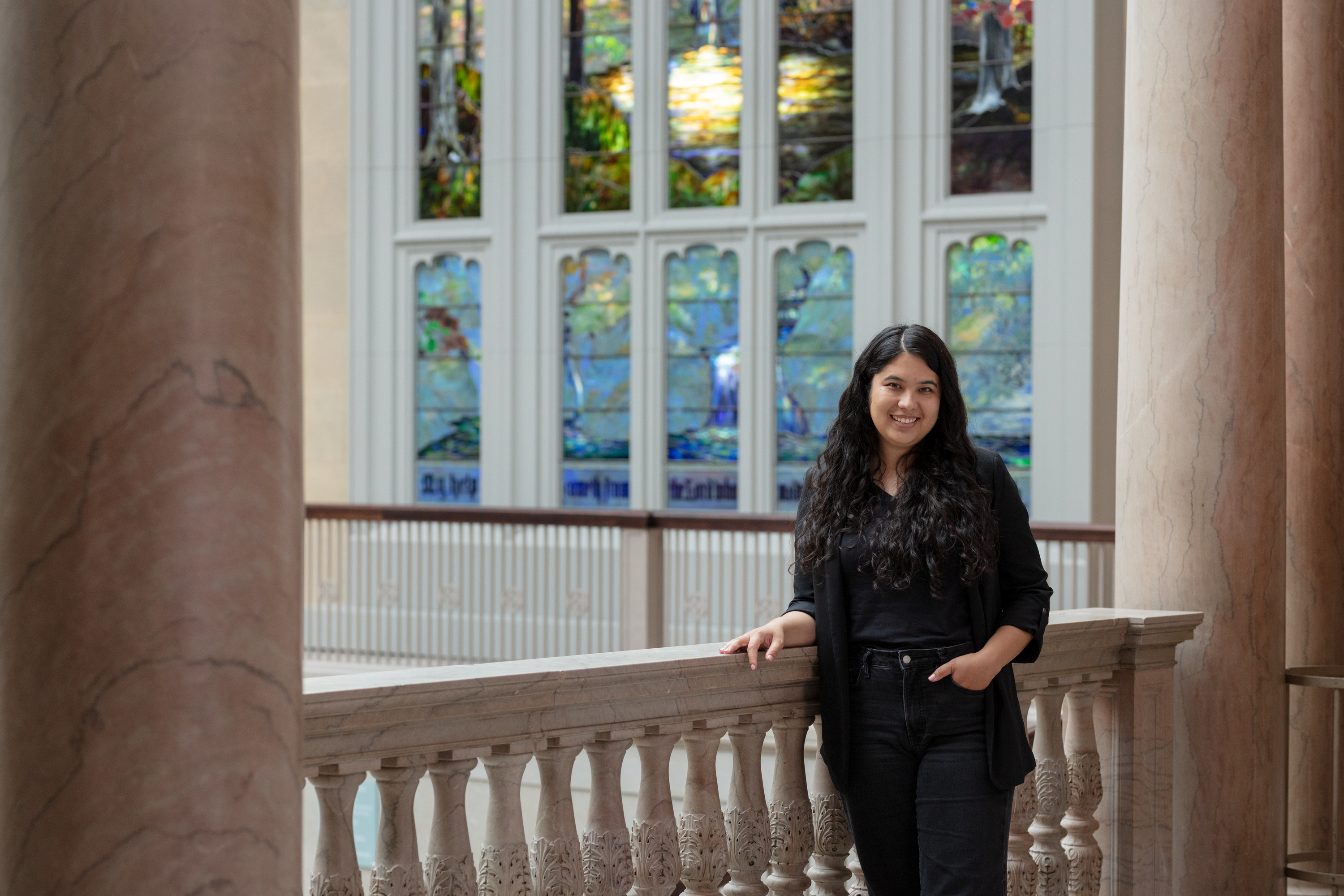 Photo of a young woman with medium-light skin and dark, long hair in curls, Kylie Escudero, standing with her arm on a railing, a beautiful stained-glass window of a waterfall behind her. She smiles and looks at the viewer.