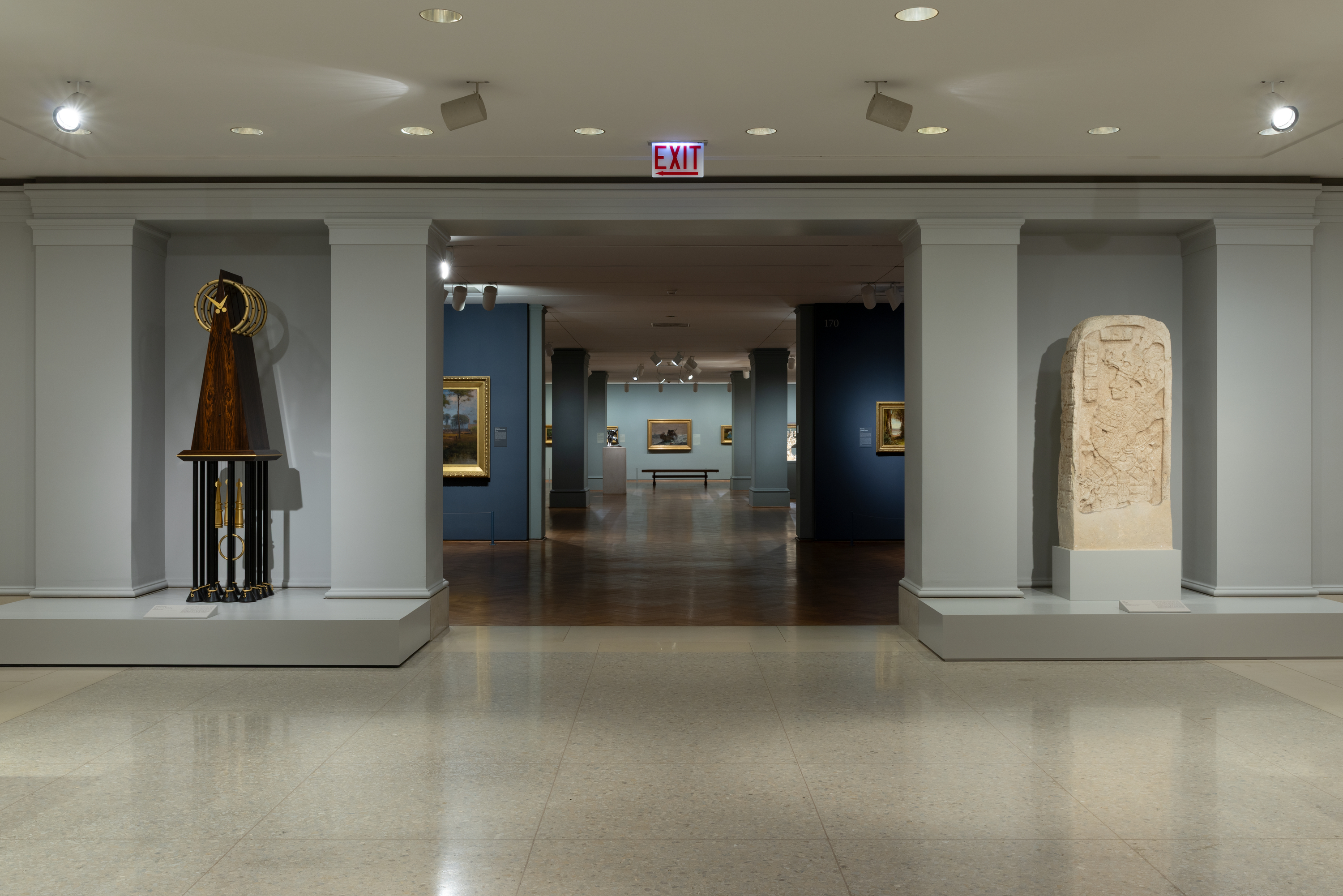 A color photograph shows a museum interior: at center a wide doorway leads to a long gallery with blue walls, framed paintings, and a bench, at left a tall dark-wood cone sculpture on a black stand, at right a pale stone carved slab, and an EXIT sign hangs above.