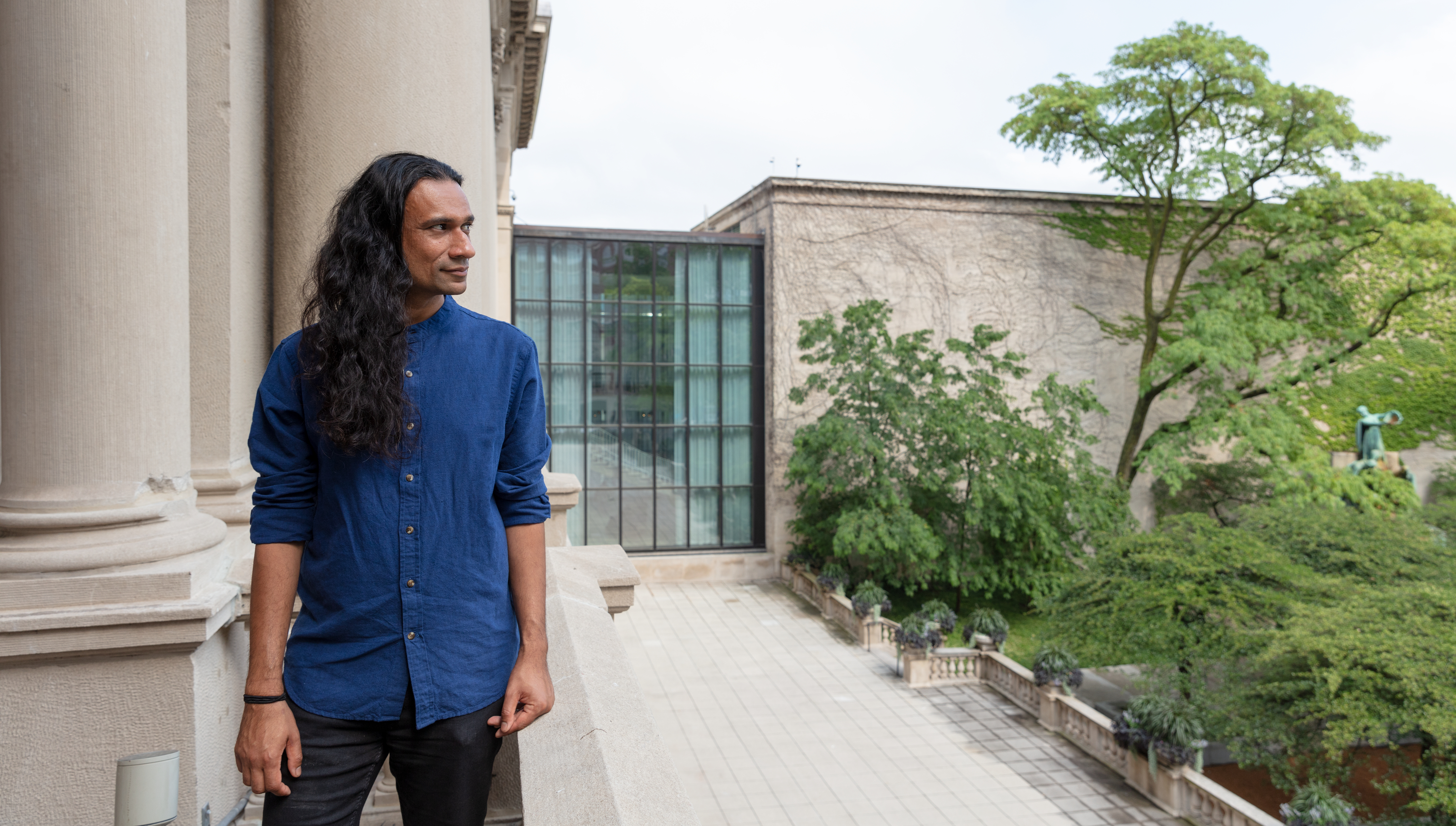 Photograph of Juneer Kibria, a medium-skinned man with long, wavy black hair, standing on a concrete balcony and staring intensely to his left toward the greenery beyond.