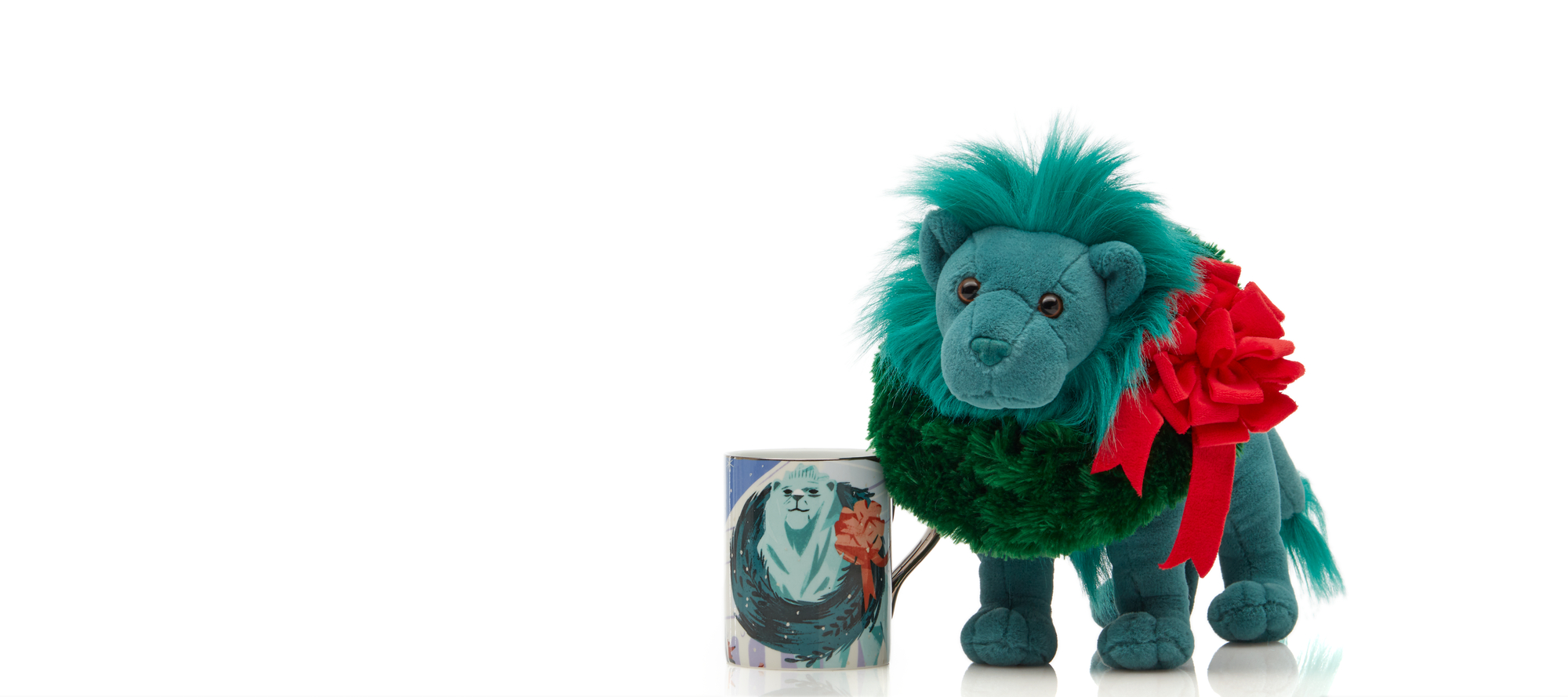 A teal plush lion with an evergreen plush wreath around its neck sits next to a white mug with an illustration of a similarly green and wreathed lion on it.