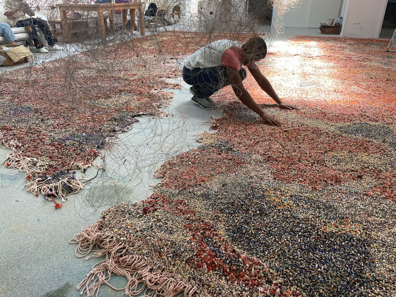 A young medium-skinned man squats as he puts his hands down on an irregularly shaped woven work on the concrete floor of an interior space.