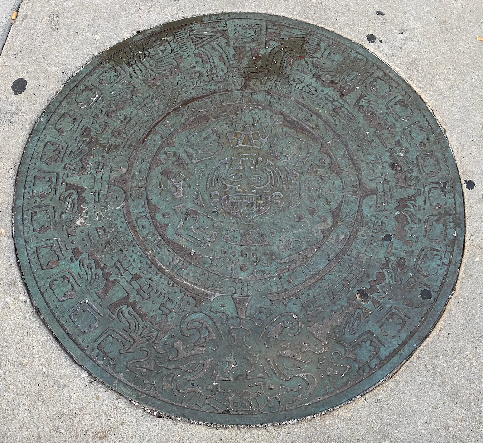 A color photograph shows a circular metal cover set in light gray pavement; at center is a face-like design, encircled by concentric rings of intricate geometric motifs, with a worn greenish-brown patina and raised patterns reaching the rim.