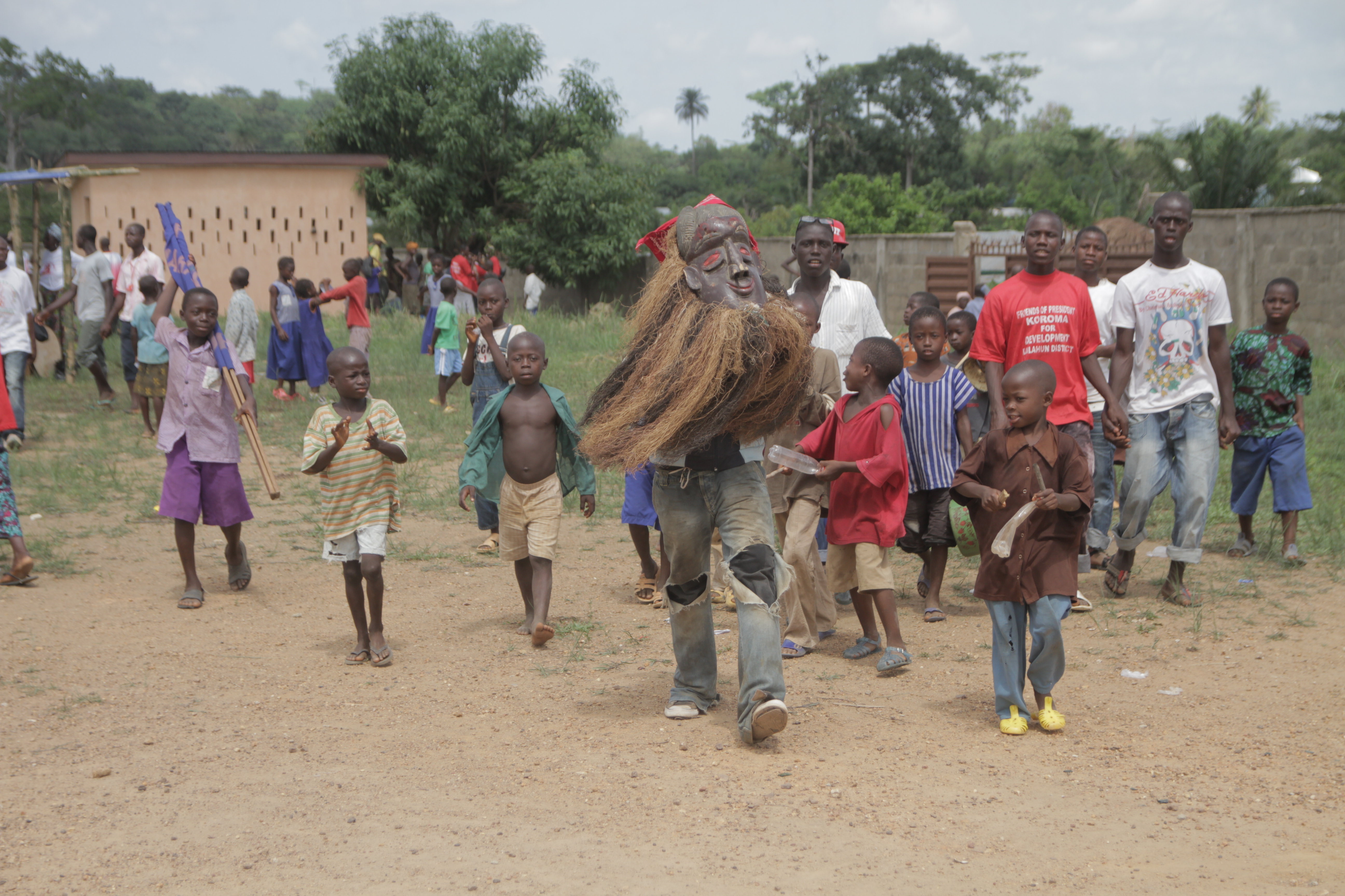 A group of black children and teens walk behind a masked figure wearing ripped jeans. The mask is dark brown, smiling, and red eyed, with a prominent brow. It is encircled with a natural fiber that forms a light-brown lion's mane that goes down to the wearer's hips.