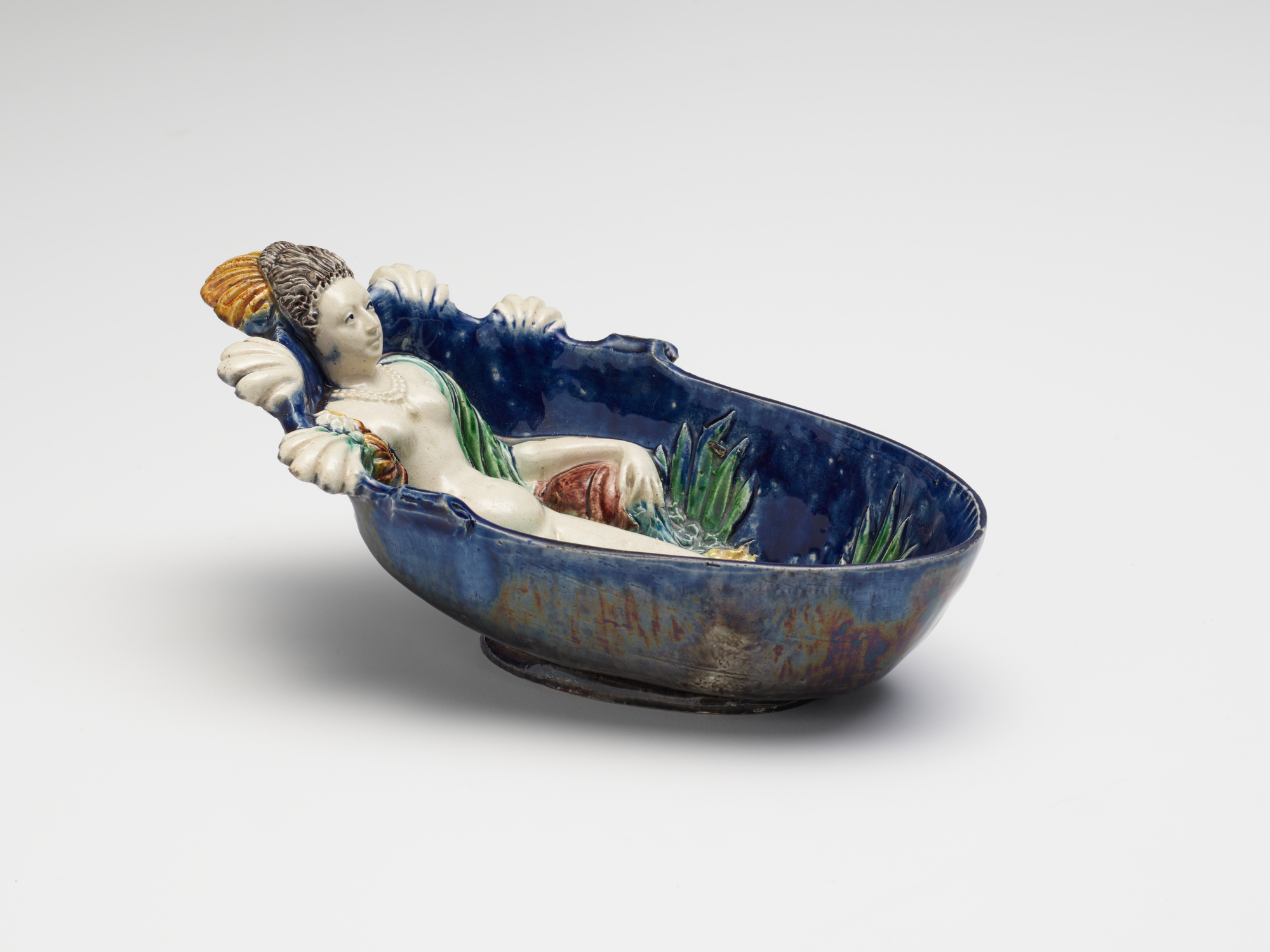 A color photograph shows a glazed ceramic bowl featuring a sculpted reclining adult figure with light skin, brown and yellow hair, and green drapery; at left the hands grasp the rim, at center the body lies among green leaves, and at right the deep blue wall curves outward.