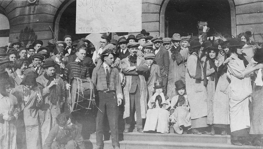 At the top center, a large hand-lettered sign reads “SEPTEMBER MORN” with “MEGA SHOW” below, hanging on a stone building with arched doorways. Across the middle, a dense crowd of adults stands on the steps in black, white, and gray tones, many wearing hats and long coats; near center-left a person holds a large drum, and another in a striped jacket stands nearby. In the foreground at the lower center and right, several people sit on the steps, including two small seated figures in light outfits and hats, while more people in long smocks and scarves line the right side.