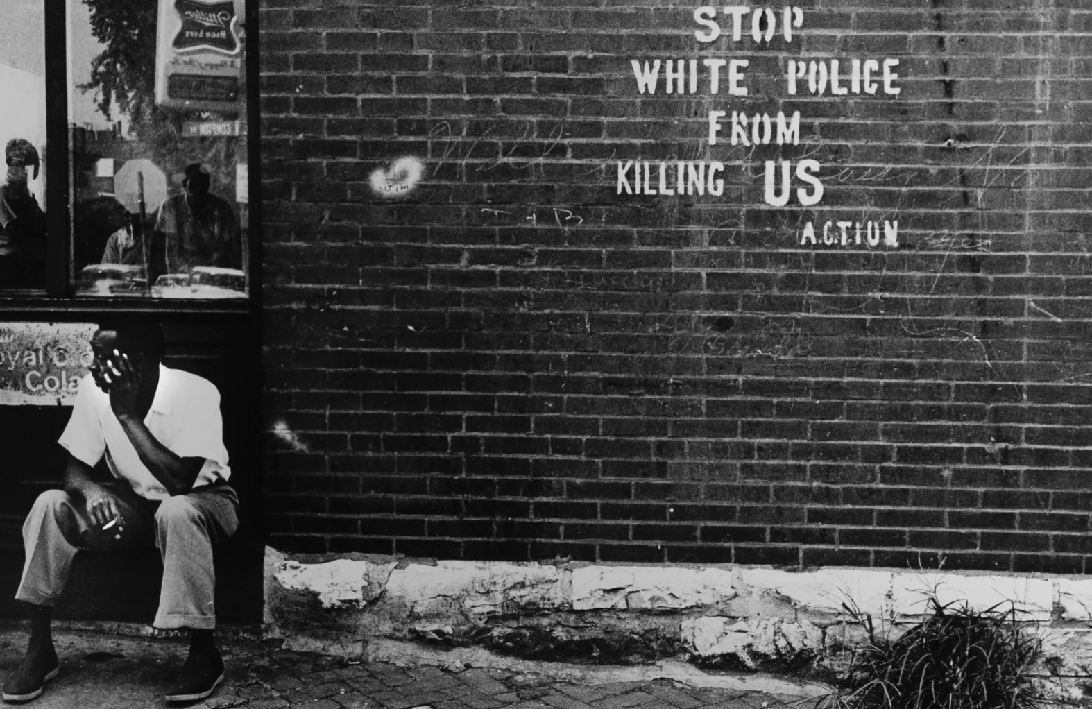 Photograph of a man seated outside against a brick wall, holding a cigarette in one hand and resting his head in the other. To the right on the wall, white block letters read: "STOP WHITE POLICE FROM KILLING US – A.C.T.I.O.N." A window on the left reflects several figures looking out from inside a building.