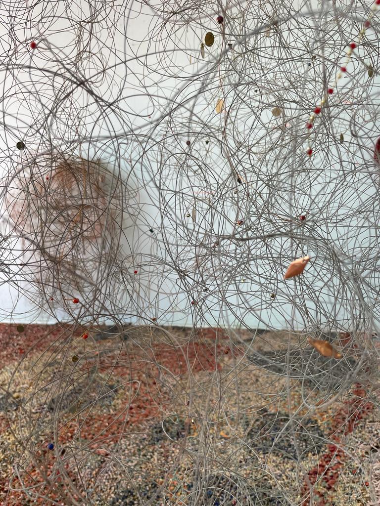 This detail focuses on the "cloud sculptures" that hang above the floor weaving. Thin silver wire is twisted and tangled, occasionally punctuated by small colorful beads.