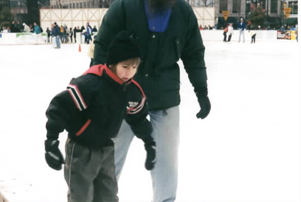 A color photograph shows a child at left in a black jacket with red stripes, gray pants, black hat and gloves beside an adult at center-right in a green coat, blue jeans, and black gloves on a white outdoor ice rink, with skaters and buildings in the background.