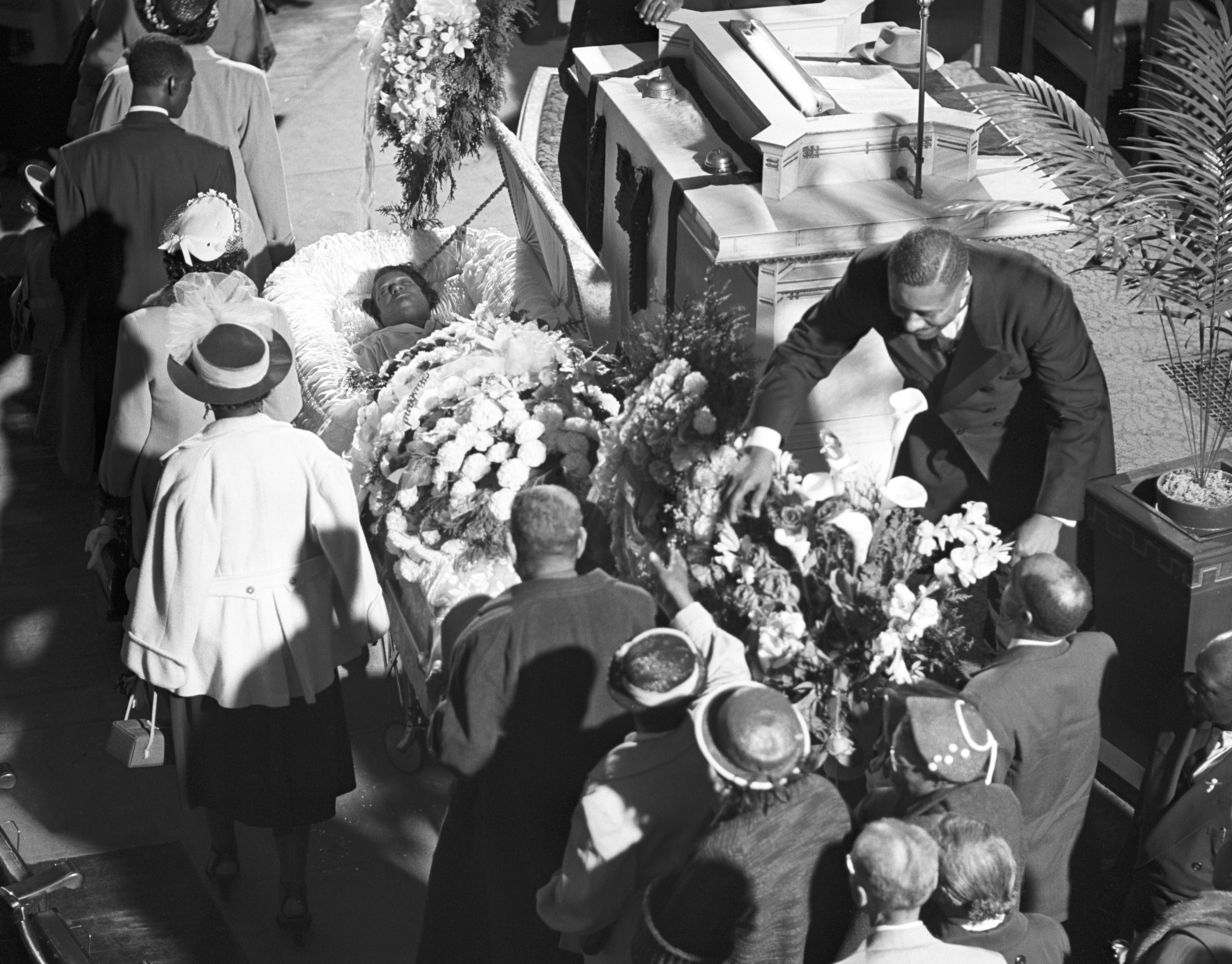 View from above of a church funeral service, with a line of mourners gathered around an open casket. A woman lies in the casket with her eyes closed, surrounded by elaborate white fabric and a large floral arrangement. At right, a man in a dark suit bends forward to adjust or place more flowers. Mourners wear formal attire, including hats and coats, and stand closely together in front of the pulpit, which is decorated with potted palms and more floral tributes.