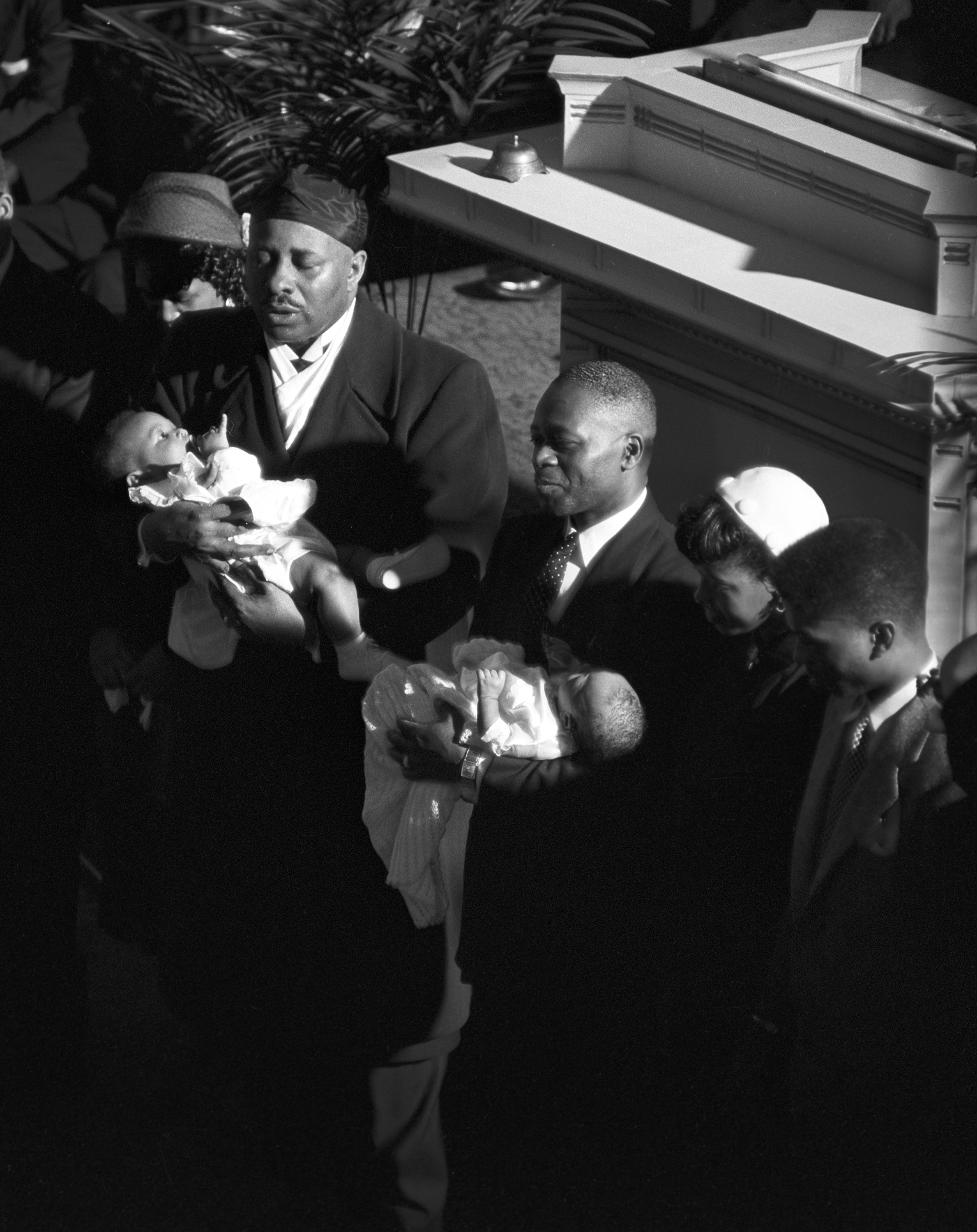 Two men in dark suits and head coverings stand near a pulpit, each cradling an infant in white clothing. A group of adults, including women in hats, stands closely around them. The scene is lit by dramatic sunlight cutting through a dark interior, likely capturing a ceremonial or religious moment such as a blessing or dedication.