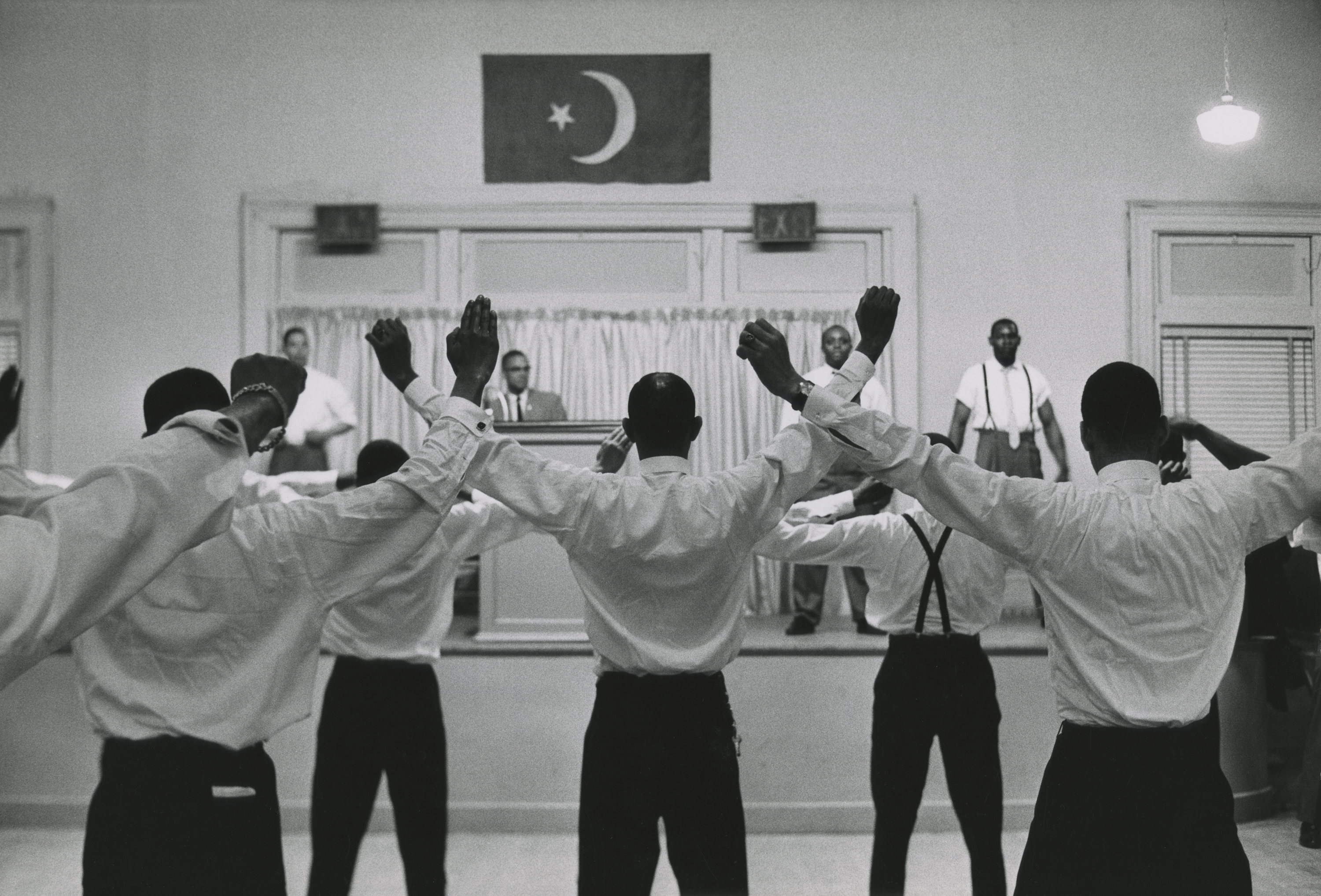 A group of men in dress shirts and slacks stand in formation, facing a small stage with their arms raised in synchronized motion. Several men onstage observe or instruct, including a man in a suit behind a podium. Above the stage hangs a flag with a crescent moon and star. The setting appears to be a meeting or training session, with bright overhead lighting and simple interior decor.