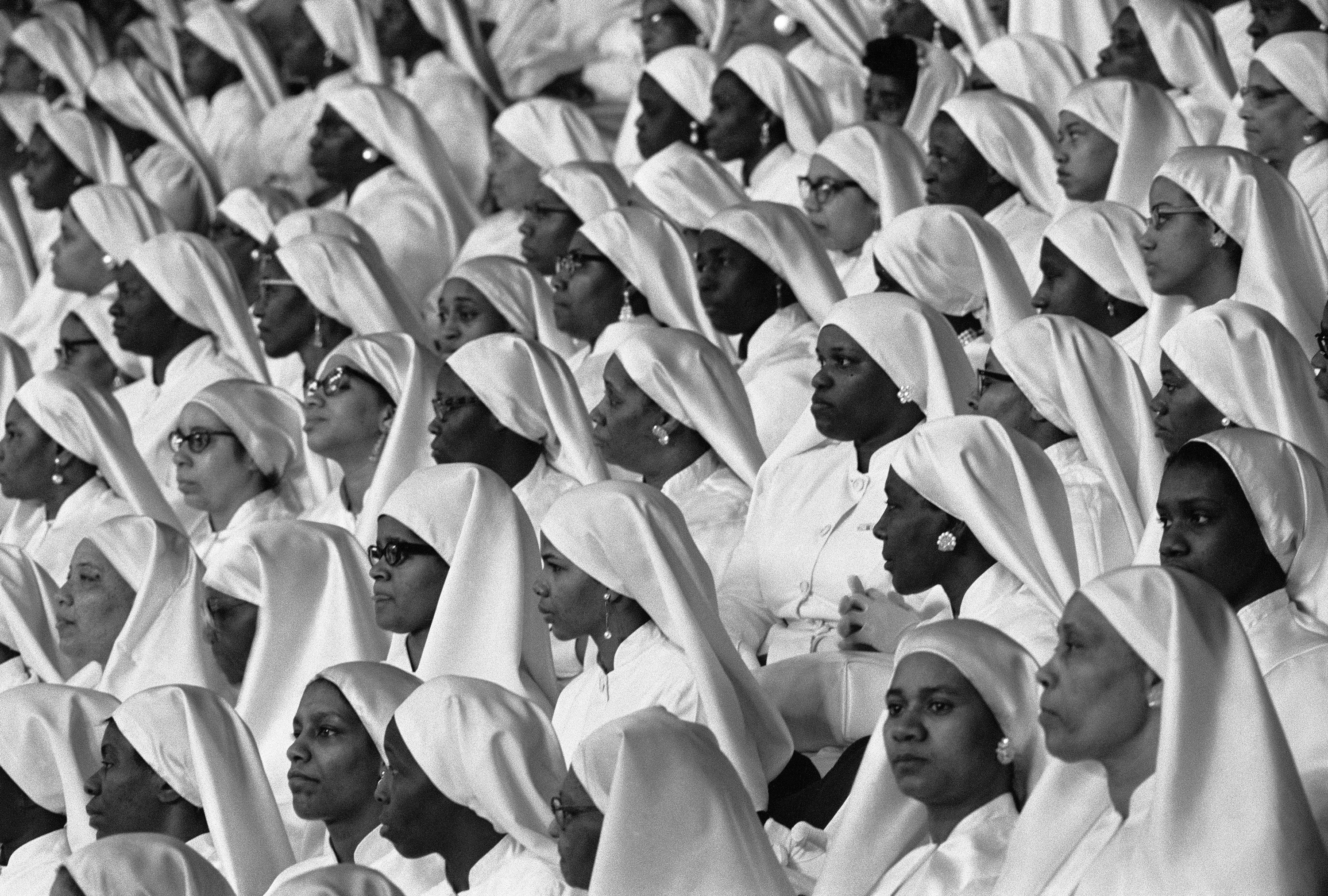 Black-and-white photograph of a large group of women seated in tightly packed rows, all dressed in matching white garments and head coverings. The women face forward with solemn or attentive expressions, many wearing eyeglasses and pearl earrings. The head coverings drape over their shoulders in a uniform style, creating a rhythmic visual pattern across the composition. The image captures a sense of order, dignity, and shared purpose among the group.