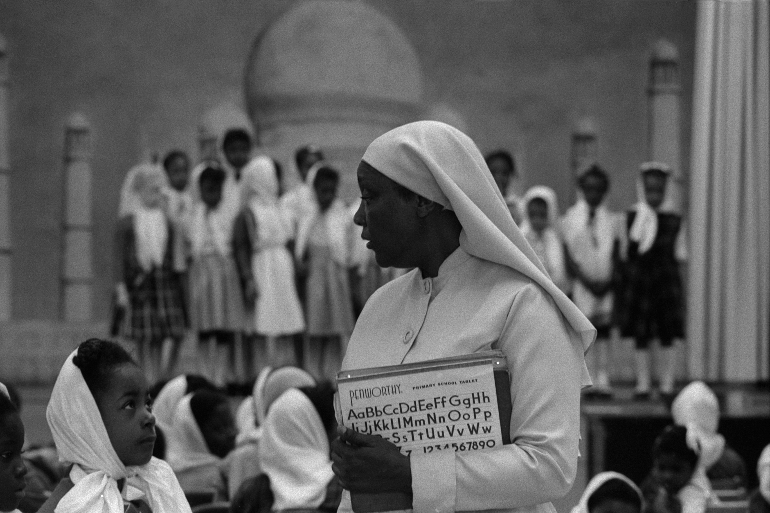 A woman wearing a white buttoned garment and head covering stands in the foreground, holding a primary school tablet with printed letters and numbers. She looks to her left toward a group of girls, all wearing headscarves and dresses, seated in rows and standing on risers in the background. A young girl in the lower left corner looks up at the woman.