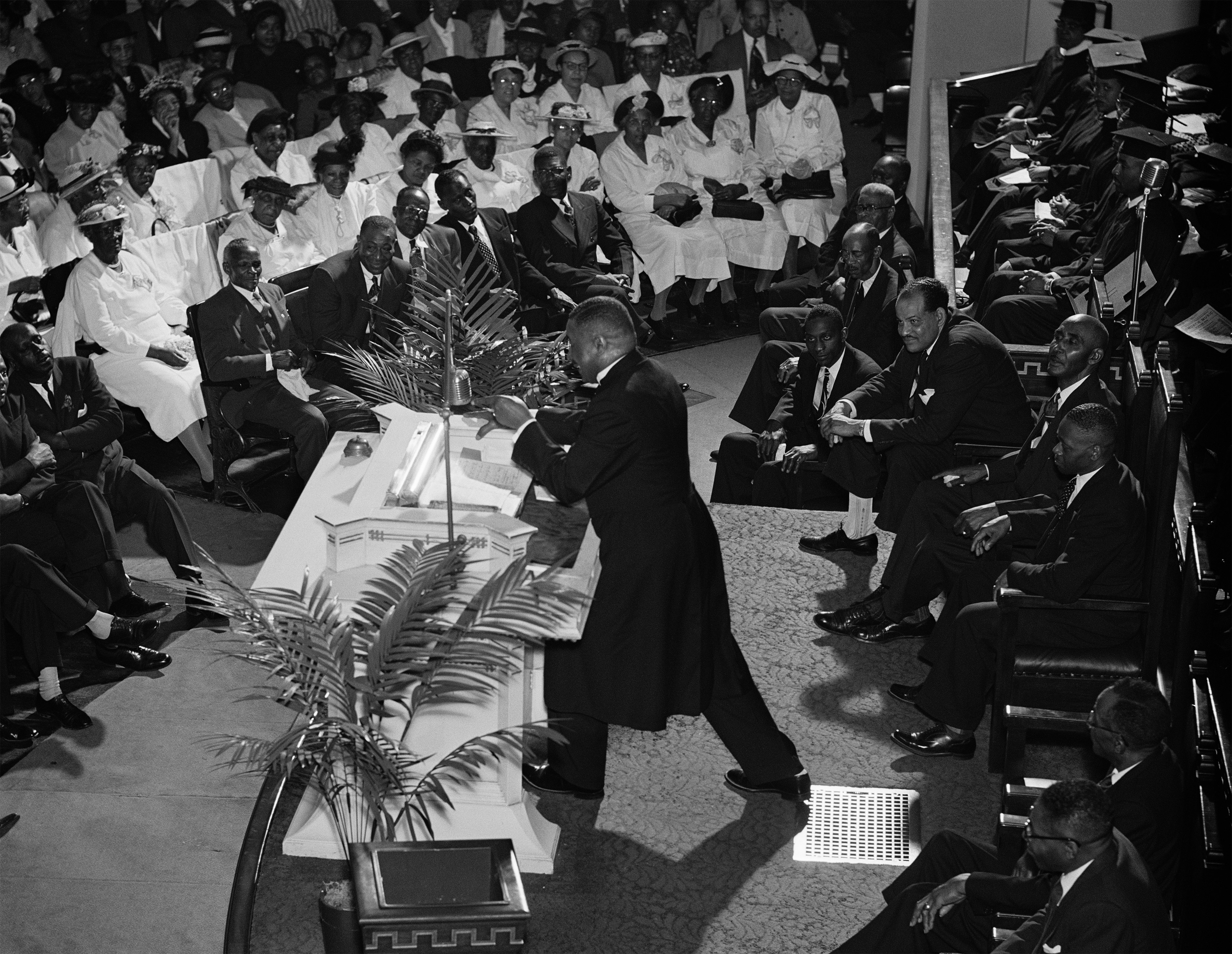 A preacher in a long black robe stands at the pulpit, leaning forward with one foot raised as he gestures emphatically. The sanctuary is full, with rows of seated men in suits and women in white dresses and hats. A group of seated men in academic robes are gathered to the preacher’s right, and potted palms decorate the pulpit area.