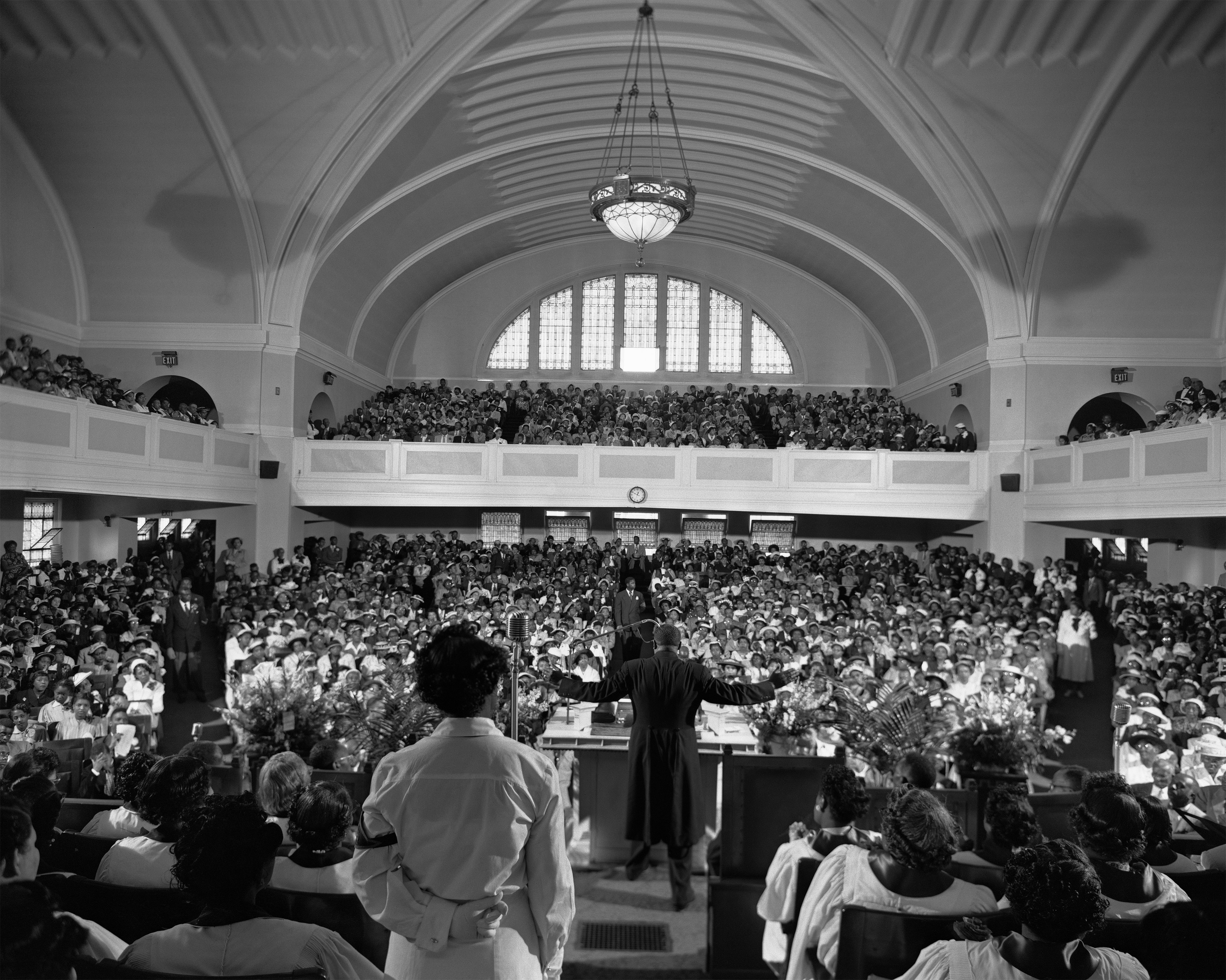 A preacher in a long black robe stands at the pulpit of a large church sanctuary, facing a packed congregation. He gestures with arms extended wide. A woman in white with her hands clasped behind her back stands at the front facing the preacher. The sanctuary is filled with men and women in formal attire, seated on the ground floor and in balconies beneath a high arched ceiling with a large central chandelier.